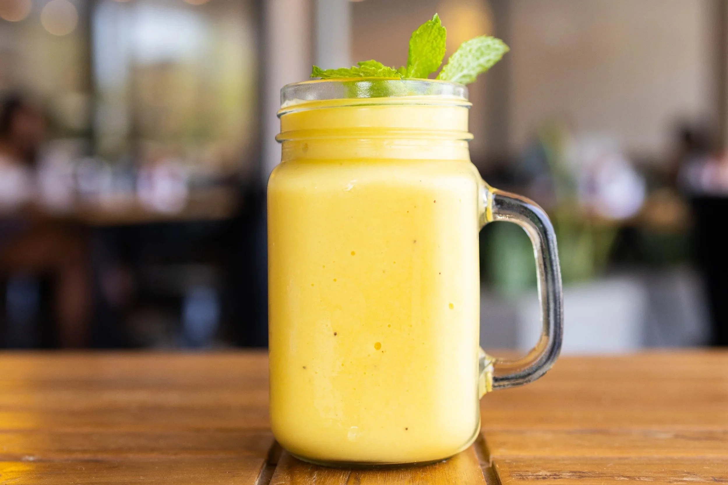 A yellow smoothie or milkshake in a glass jar with a handle, garnished with mint leaves, on a wooden table.