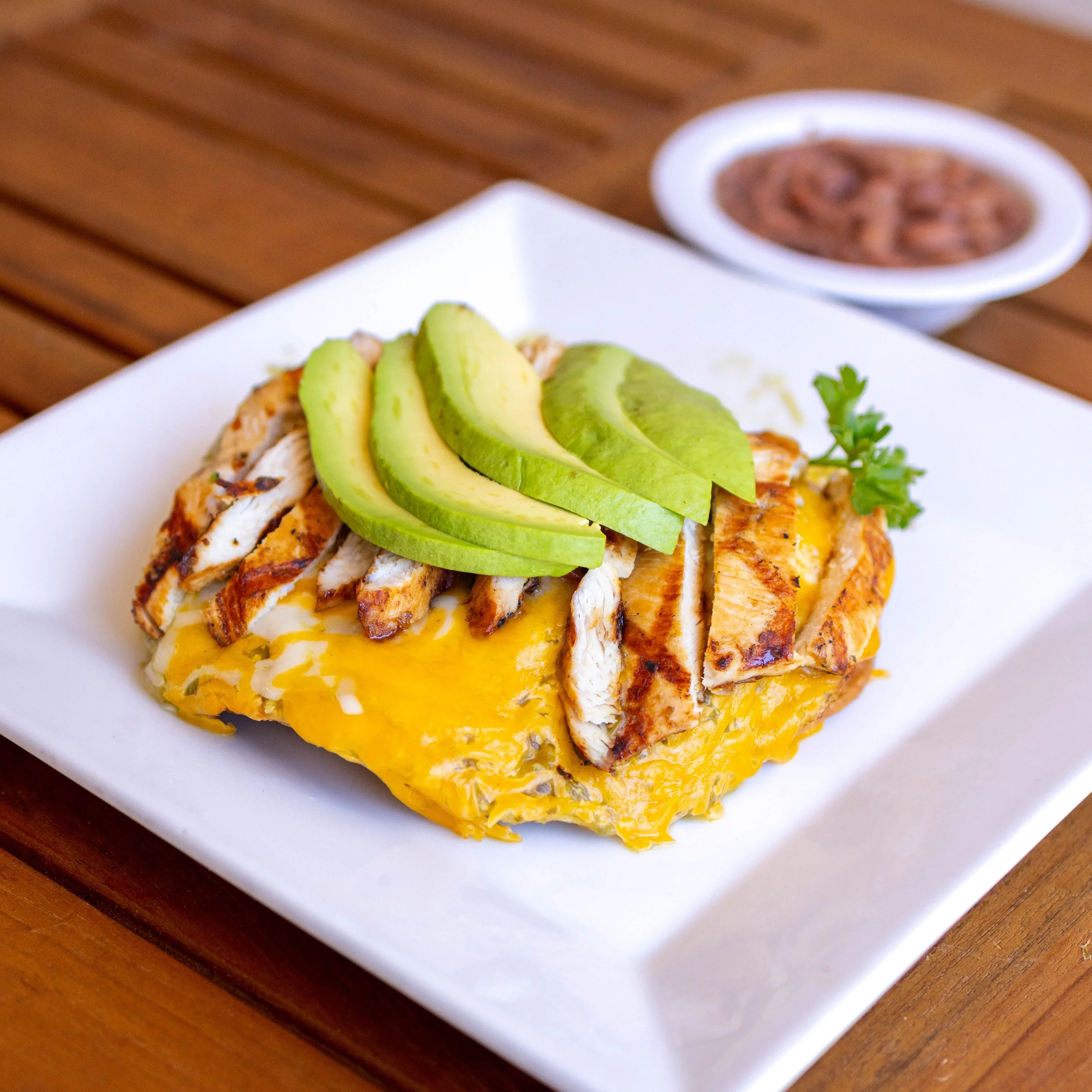 An omelette topped with grilled chicken slices, avocado slices, and a sprig of parsley, served on a white square plate with a side of salsa in a small bowl in the background.