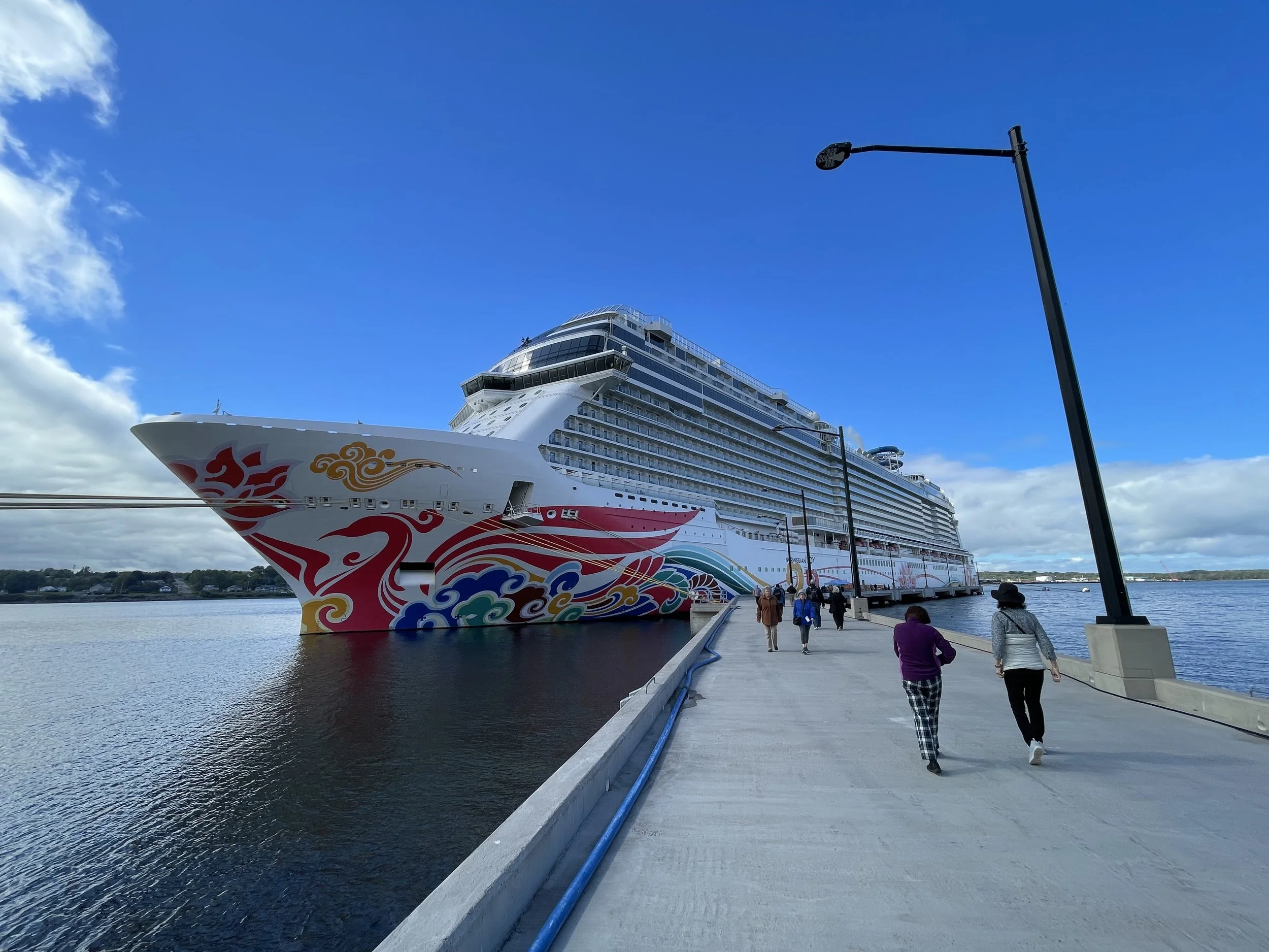 Norwegian Cruise Lines Norwegian Joy cruise ship docked at Sydney, NS, Canada pier with a colorful dragon design painted on the hull, and a few people walking along the pier under a partly cloudy sky.
