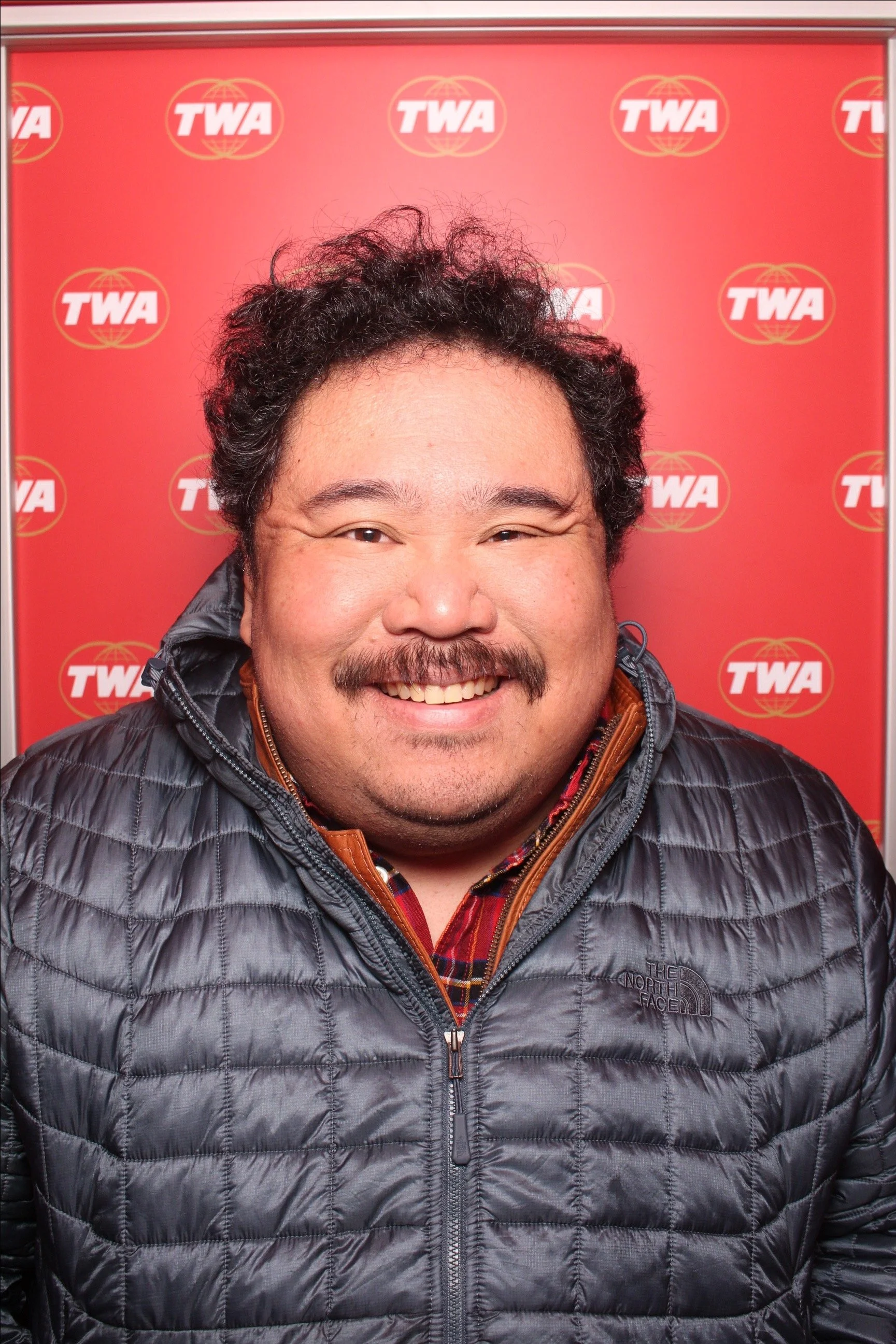 A smiling man with a mustache and curly hair wearing a black North Face puffer jacket and a plaid shirt standing in front of a red background with TWA logos.