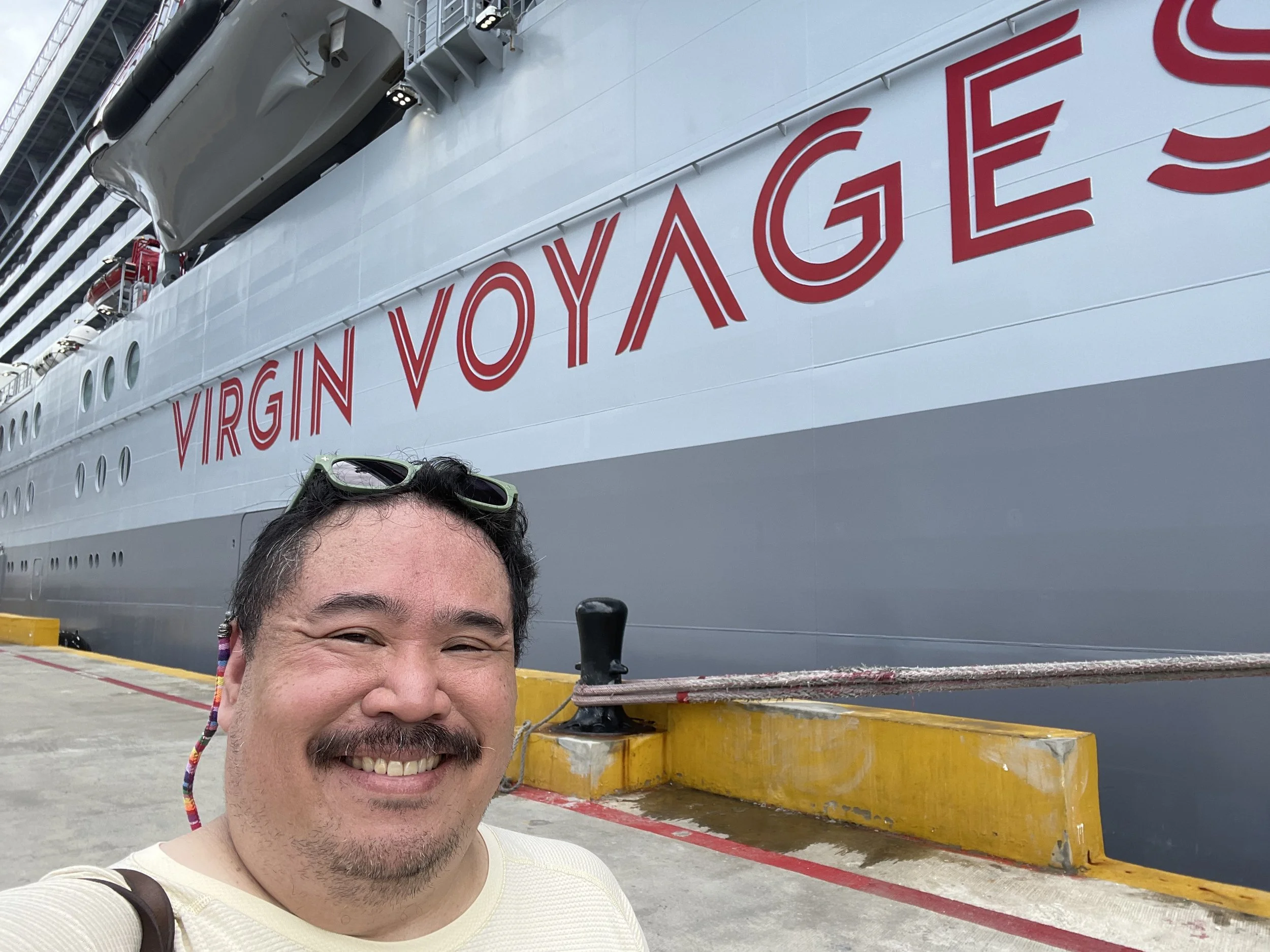 A man smiling in front of a cruise ship with red and white 'VIRGIN VOYAGES' lettering, docked at a port.