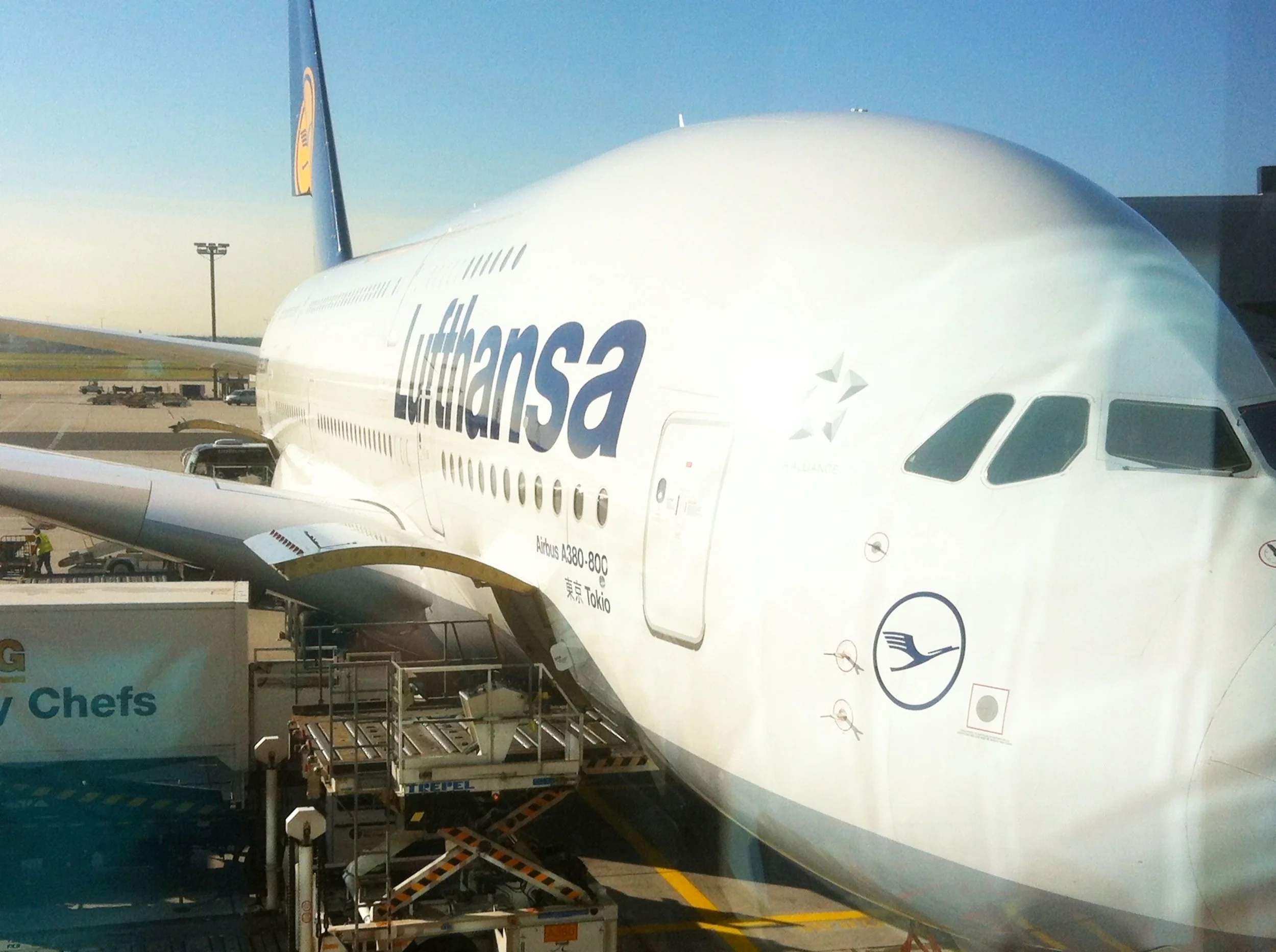 Personal pic of Lufthansa German Airlines Airbus A380 airplane at the gate at Frankfurt International Airport, connected to a jet bridge, with airport tarmac and other parked aircraft in the background during daytime.