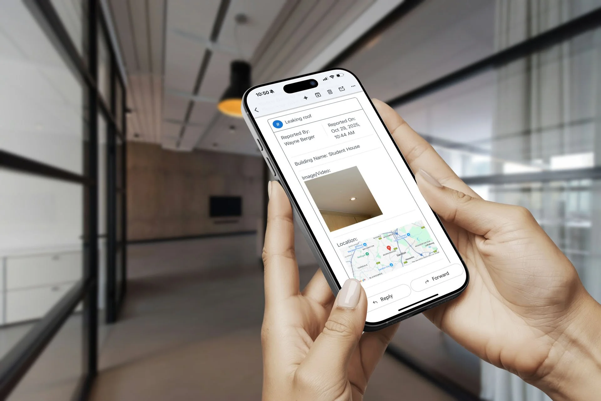 Person holding smartphone displaying a leak report about a leaking roof, with details including date, building location, an image of the ceiling, and a map, inside a modern building corridor. For Multi-location maintenance and compliance management.