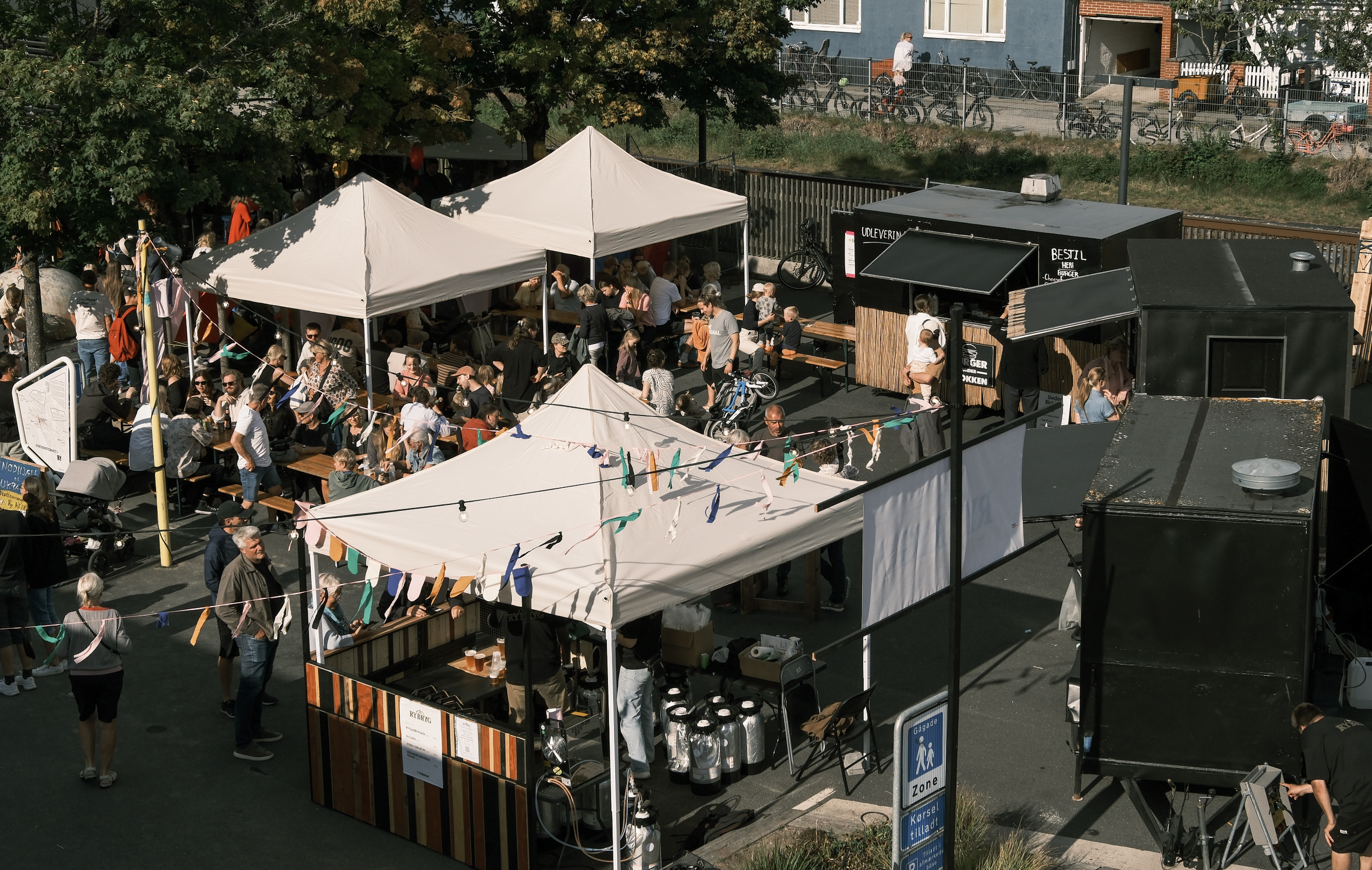 Udsigt over en udendørs markedsplads med mange mennesker, borde, telte og madboder, omgivet af træer og bygninger.