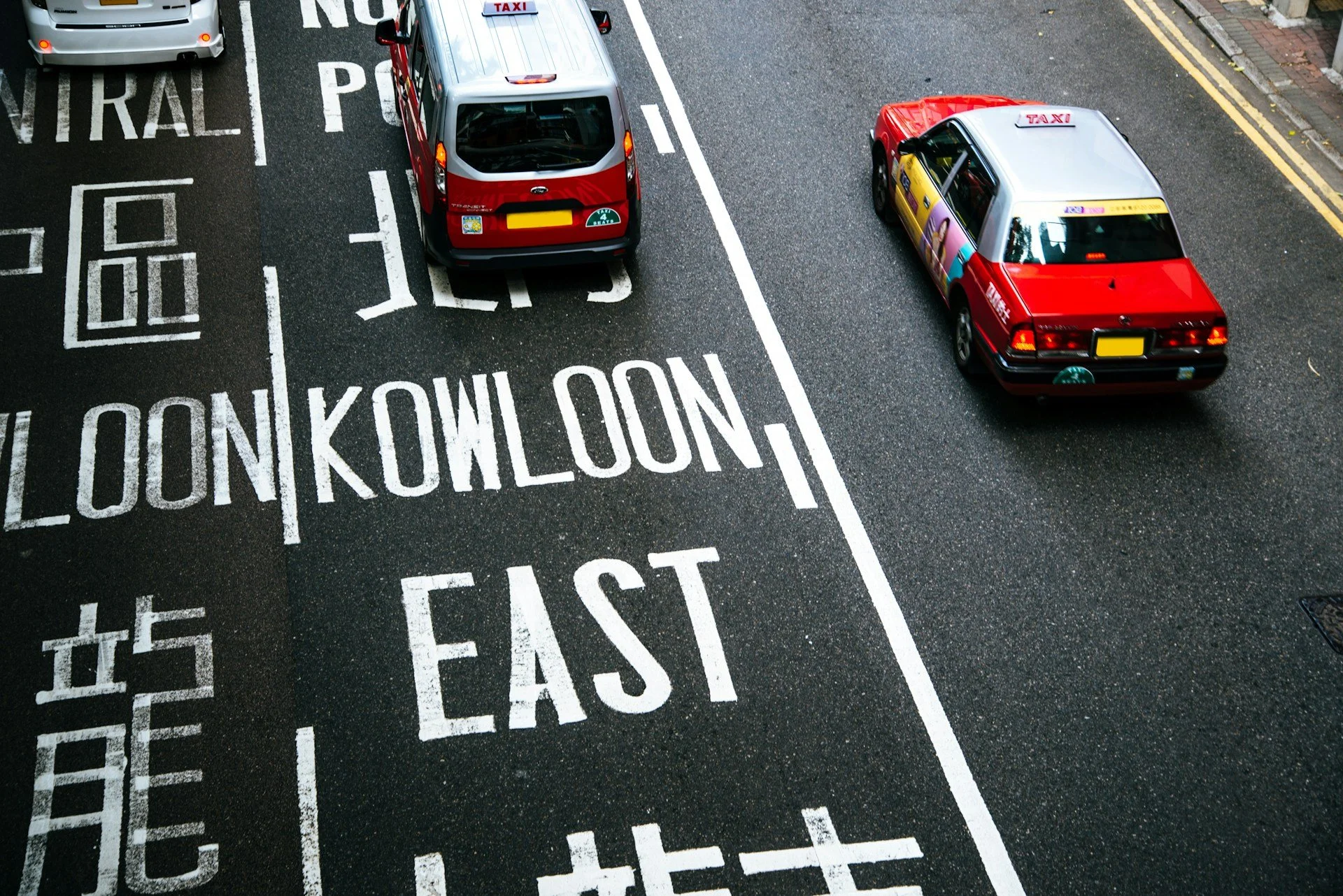 An overhead view of two taxis parked on a city street. The street has white painted lines and text, including the words 'Central' and 'East.' The taxis are red and silver, with the silver taxi featuring an advertisement on its side.