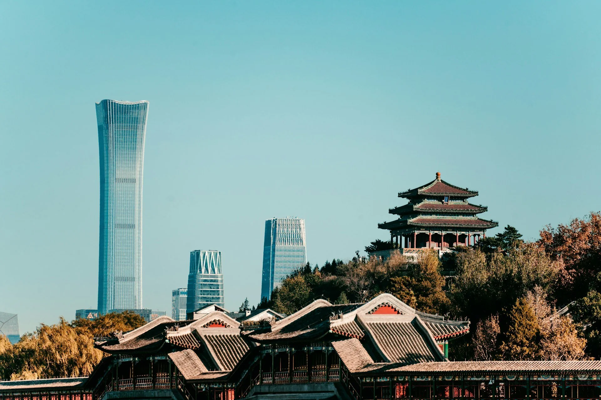 Traditional Chinese pavilion atop a hill in Beijing with modern skyscrapers in the background and trees in the foreground.