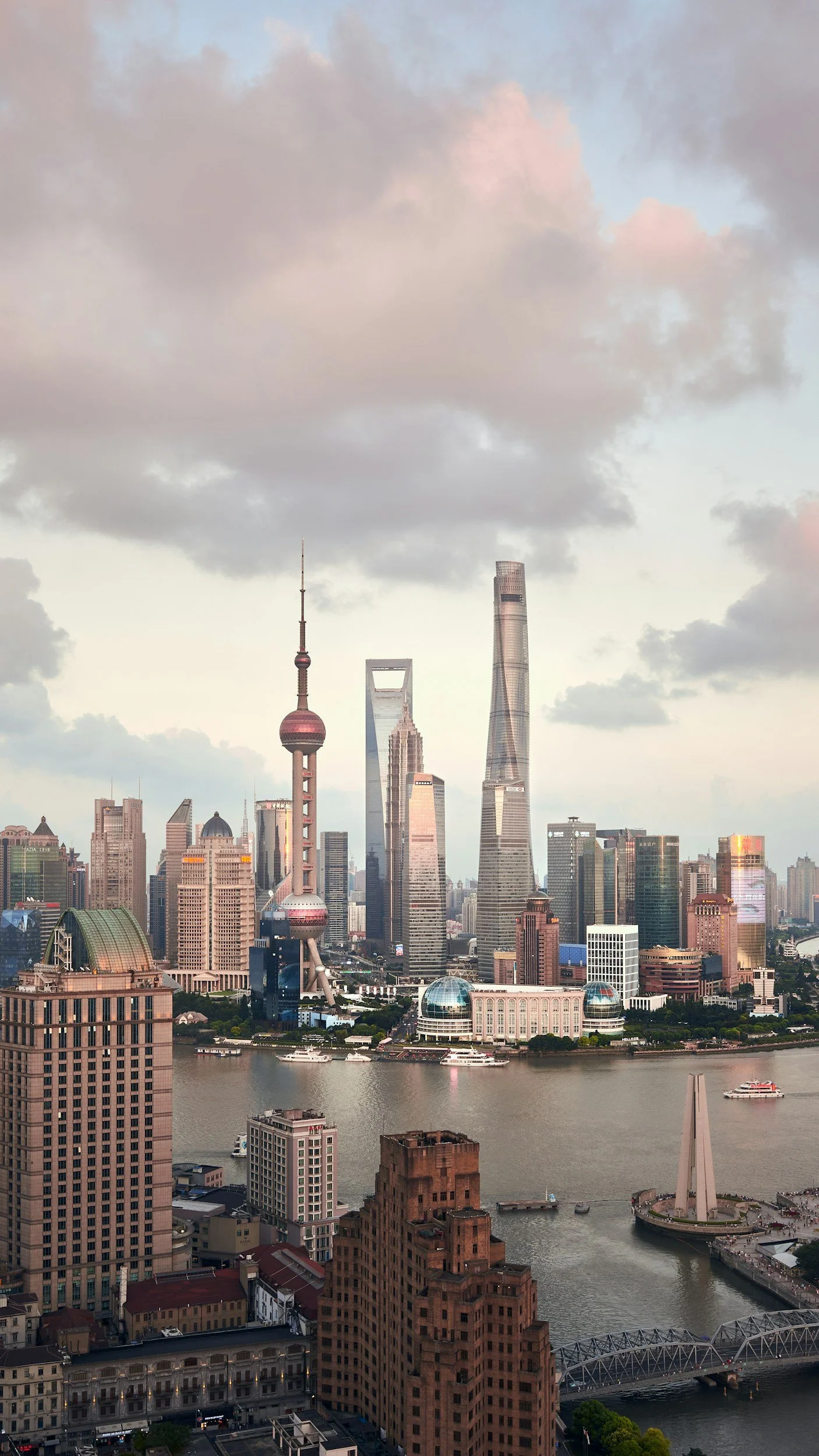 Skyline of Shanghai with the Oriental Pearl Tower, Shanghai World Financial Center, and Shanghai Tower as seen from across the Huangpu River with boats and buildings in the foreground under a cloudy sky.