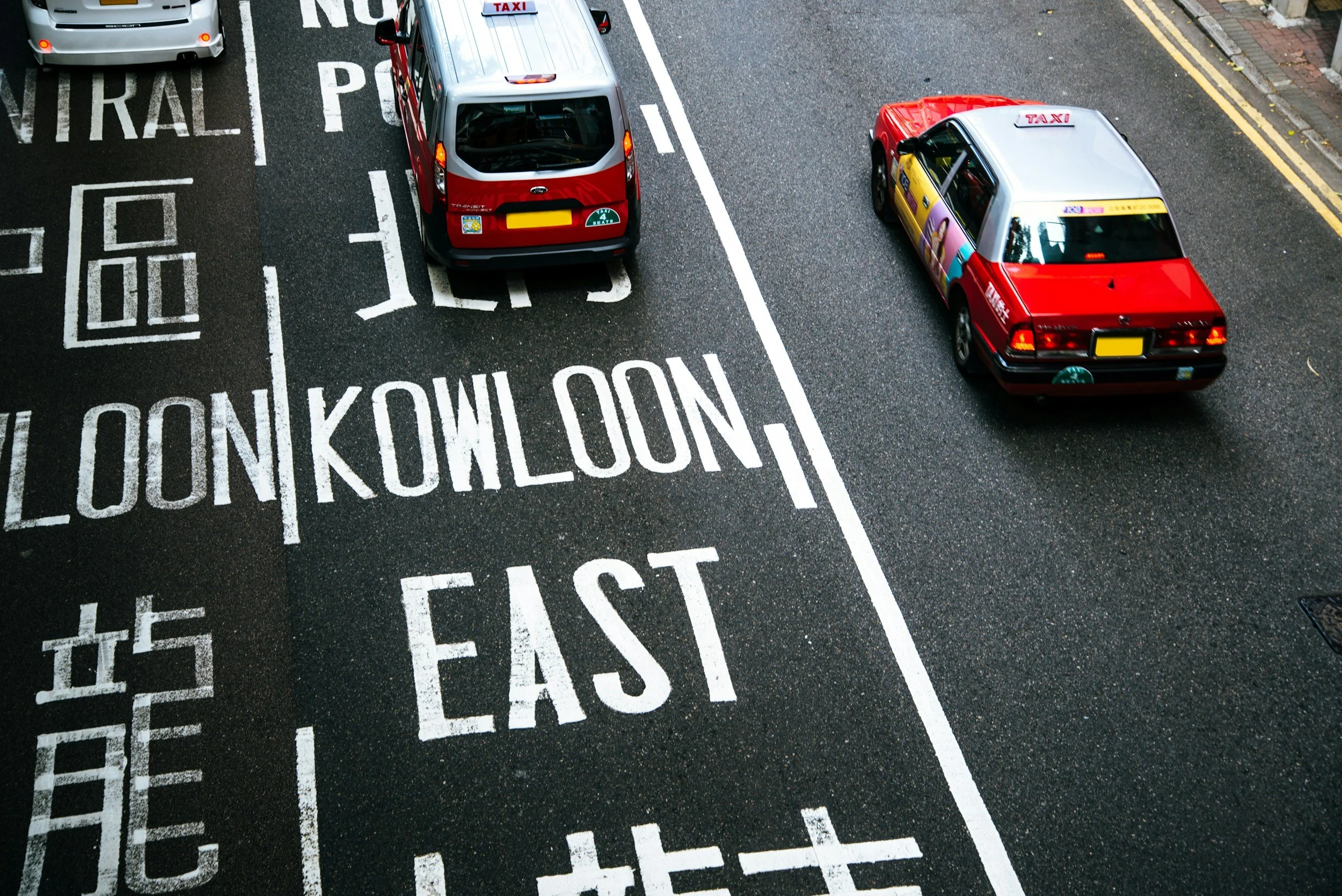 Top-down view of a city street showing painted street markings, including the words 'LOOK BOTH WAYS' and 'EAST,' with two taxis and a car parked along the curb.