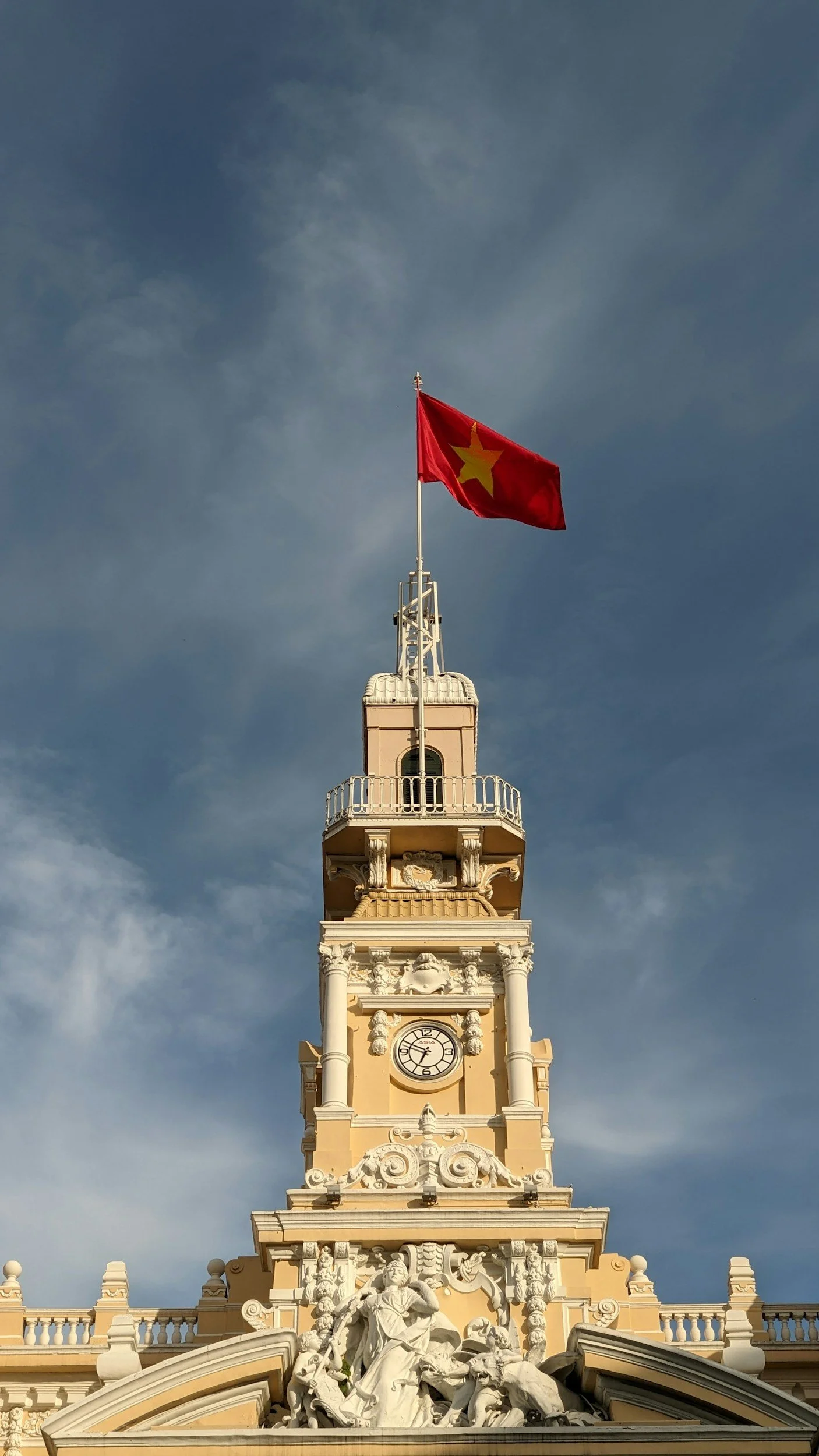 A tall ornate building with a clock and a sarcophagus top feature, with a yellow flag with a red star flying atop it against a partly cloudy sky.