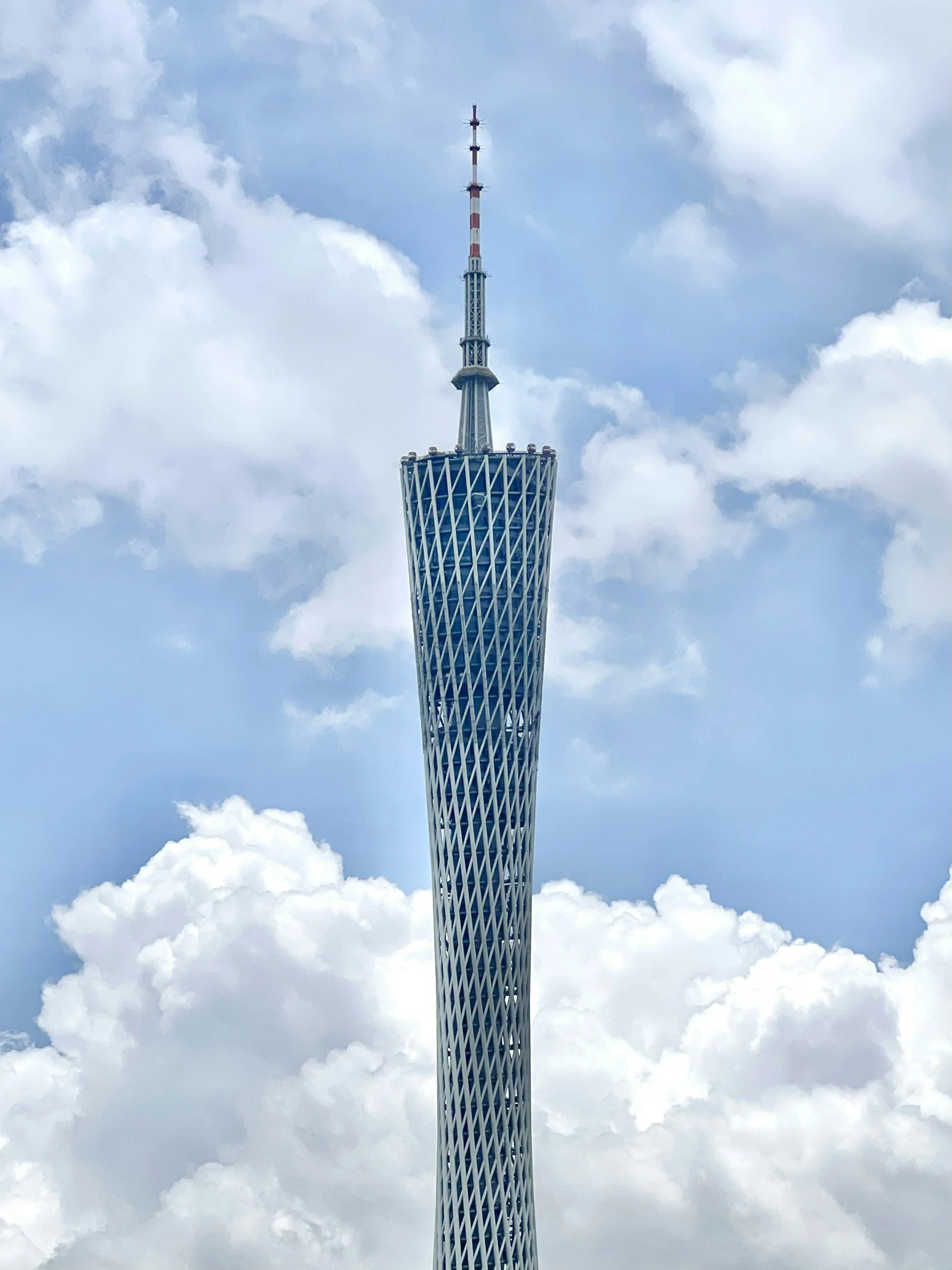 Skyline view of a tall modern skyscraper with a lattice pattern on its exterior, topped with an antenna, against a backdrop of blue sky and white clouds.