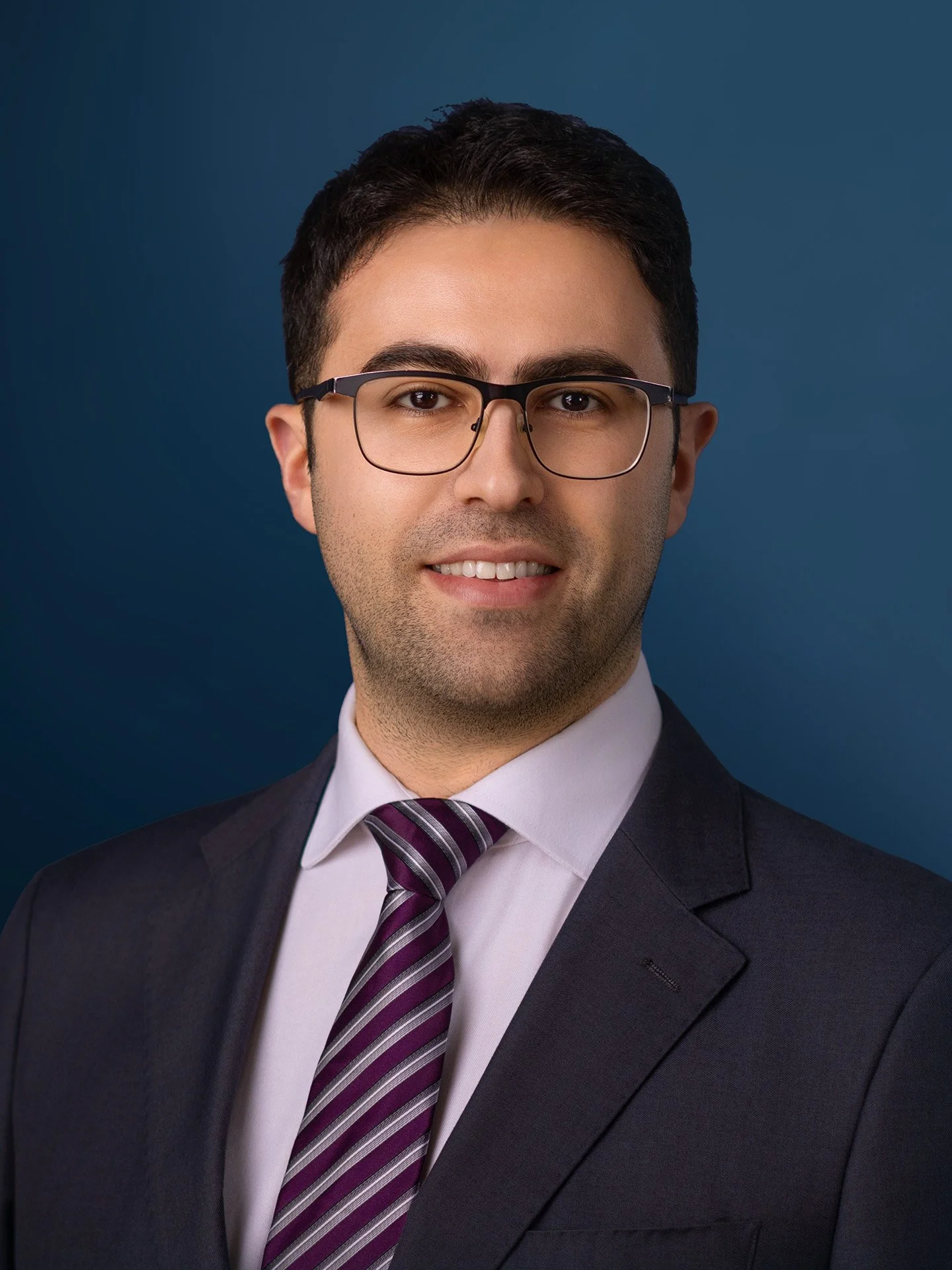 A professional headshot of a young man with dark hair, glasses, wearing a suit, white shirt, and a striped purple and gray tie, against a dark blue background.