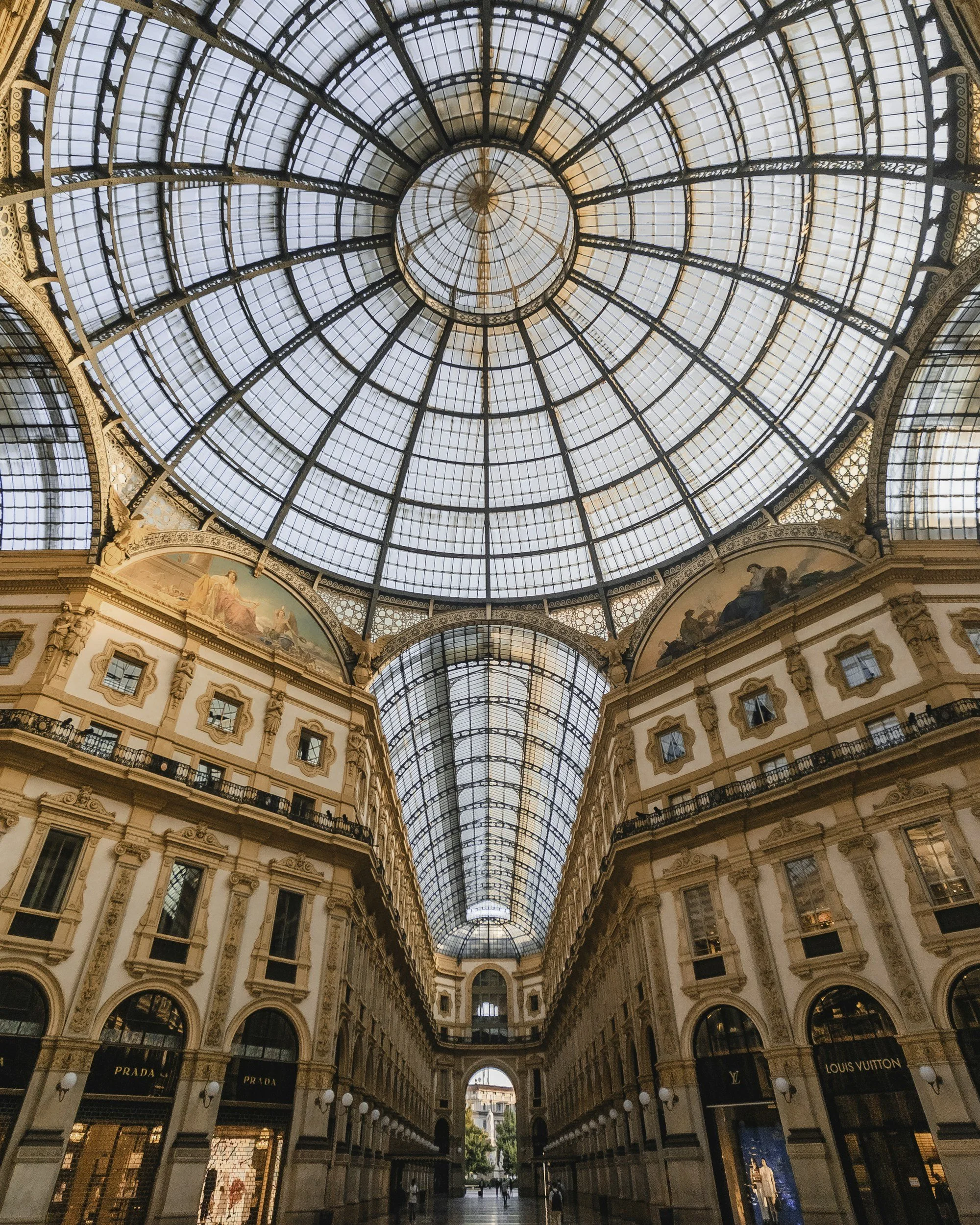 Interior view of a shopping mall with a glass-domed ceiling, classic architectural details, and luxury brand storefronts such as Prada and Louis Vuitton.