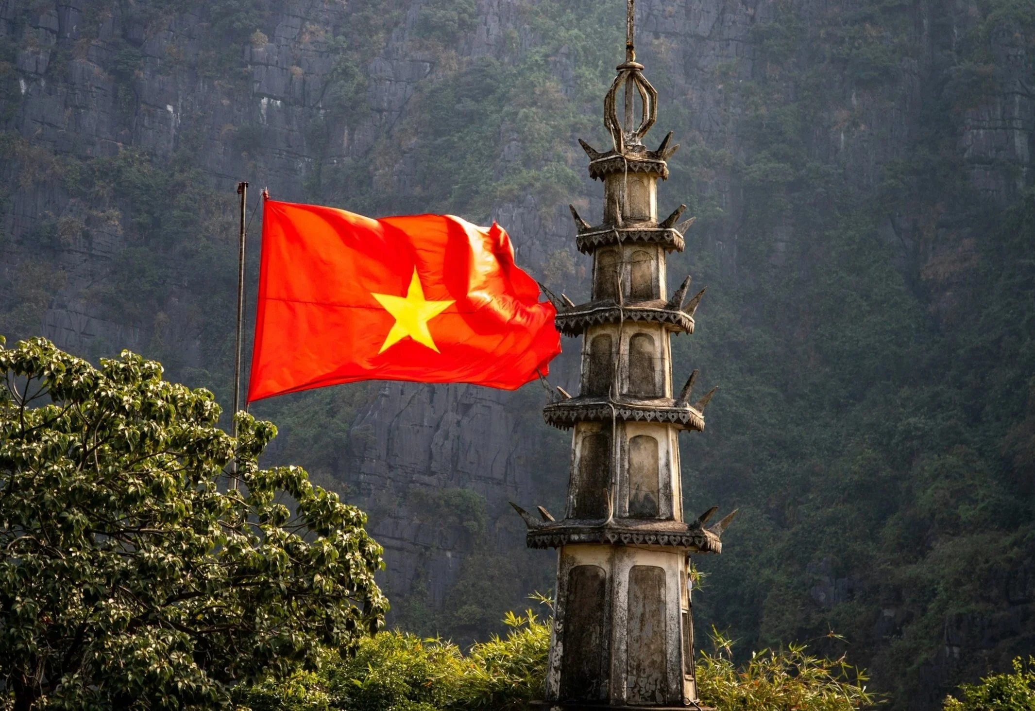 A red flag with a yellow star waving next to a tall, ornate stone pagoda against a backdrop of a mountain with trees and rocky cliffs.