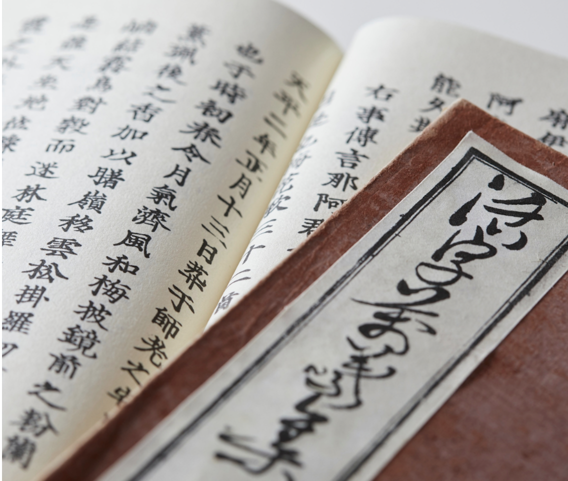 Close-up of an open book with Japanese kanji characters and a red-bordered paper with black calligraphy.