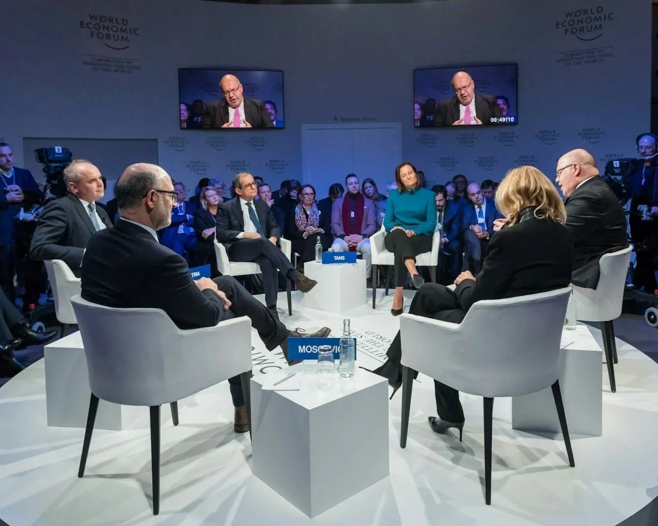A panel discussion at the World Economic Forum with six speakers sitting on white chairs in a circle, and a larger audience seated behind them. There are two TV screens showing a close-up of a man speaking, and name tags on the floor in front of some panelists labeled 'MORO,' 'TANS,' and 'MOSI.' The room has a professional and formal atmosphere.