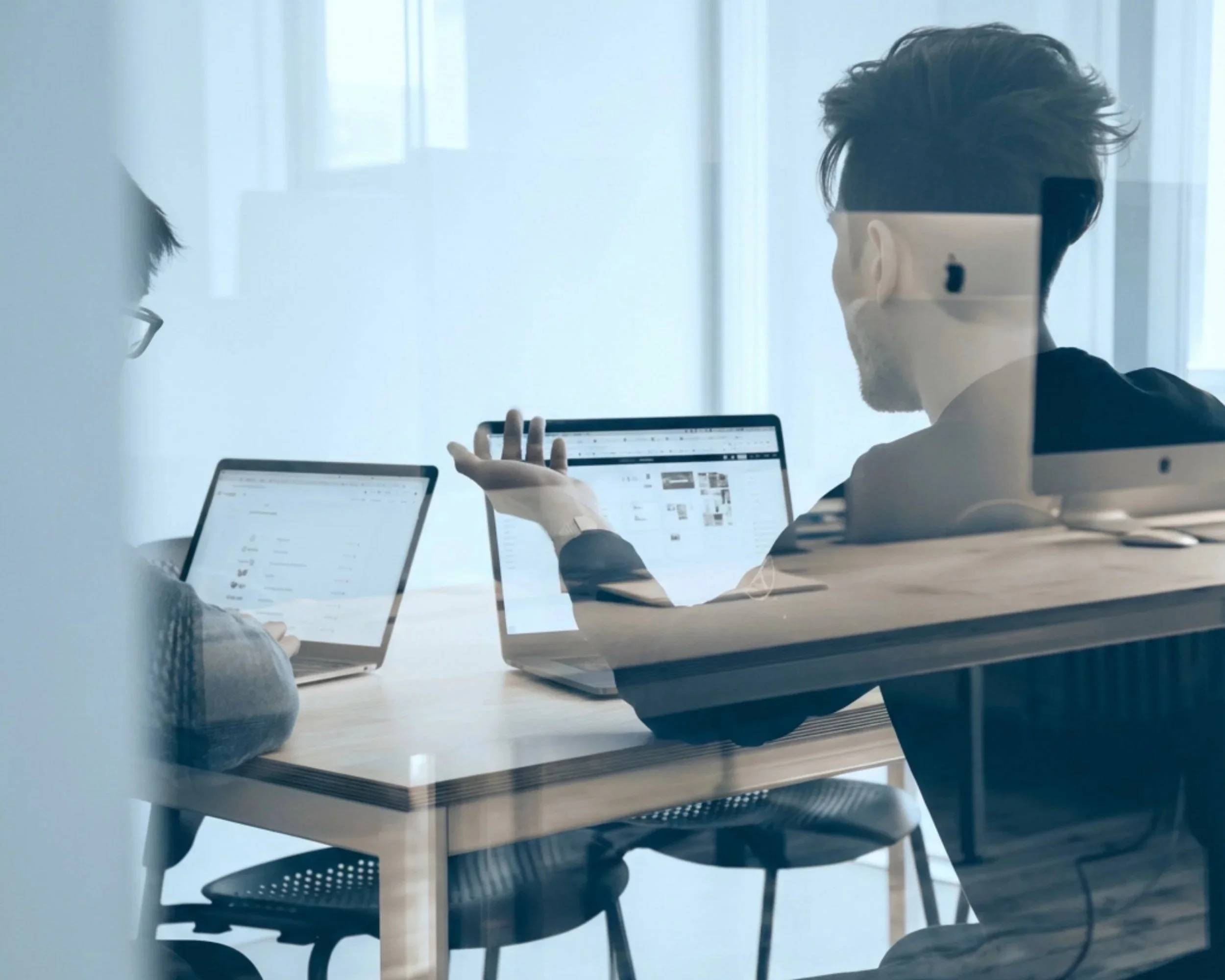 A man with styled hair wearing a face mask and black shirt, sitting at a wooden table in a modern office or conference room, gesturing with his hand towards a laptop with a webpage displayed on the screen.