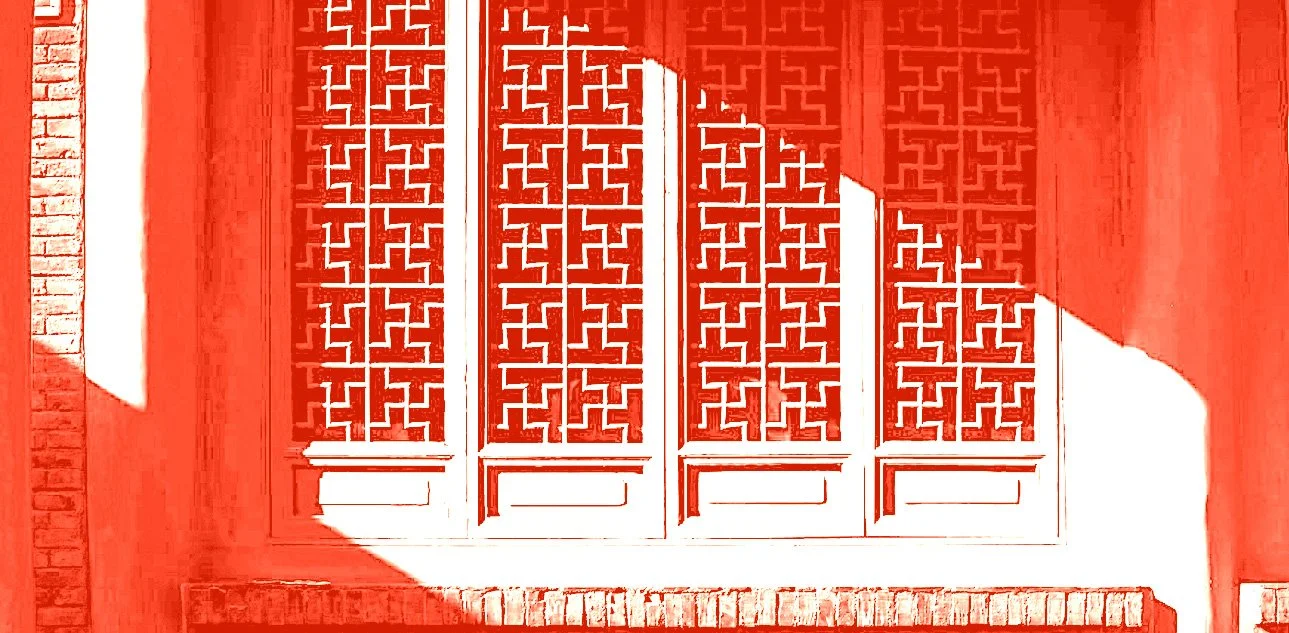 Red decorative window shutters with intricate geometric patterns on a white wall, framed by brick wall on the sides.