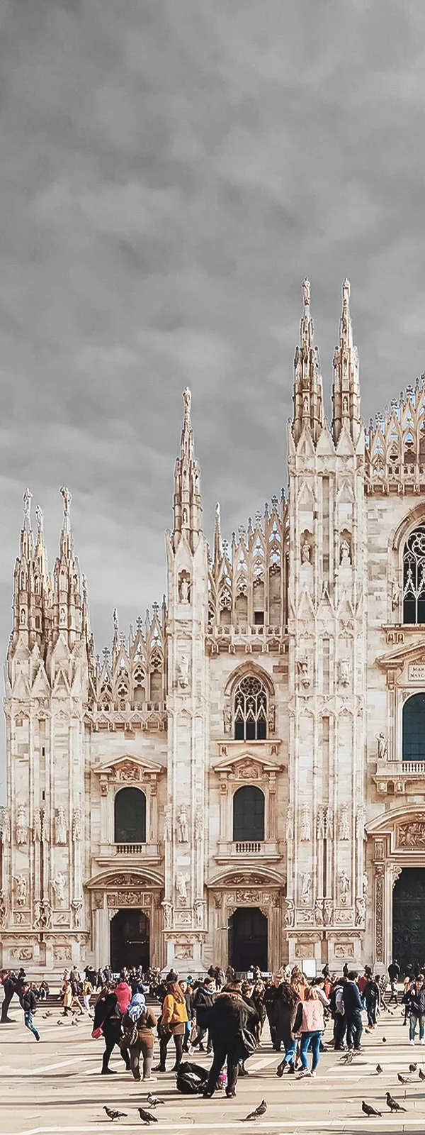 People walking and pigeons in front of Milan Cathedral, a white Gothic-style church with ornate spires and statues, under a cloudy sky.