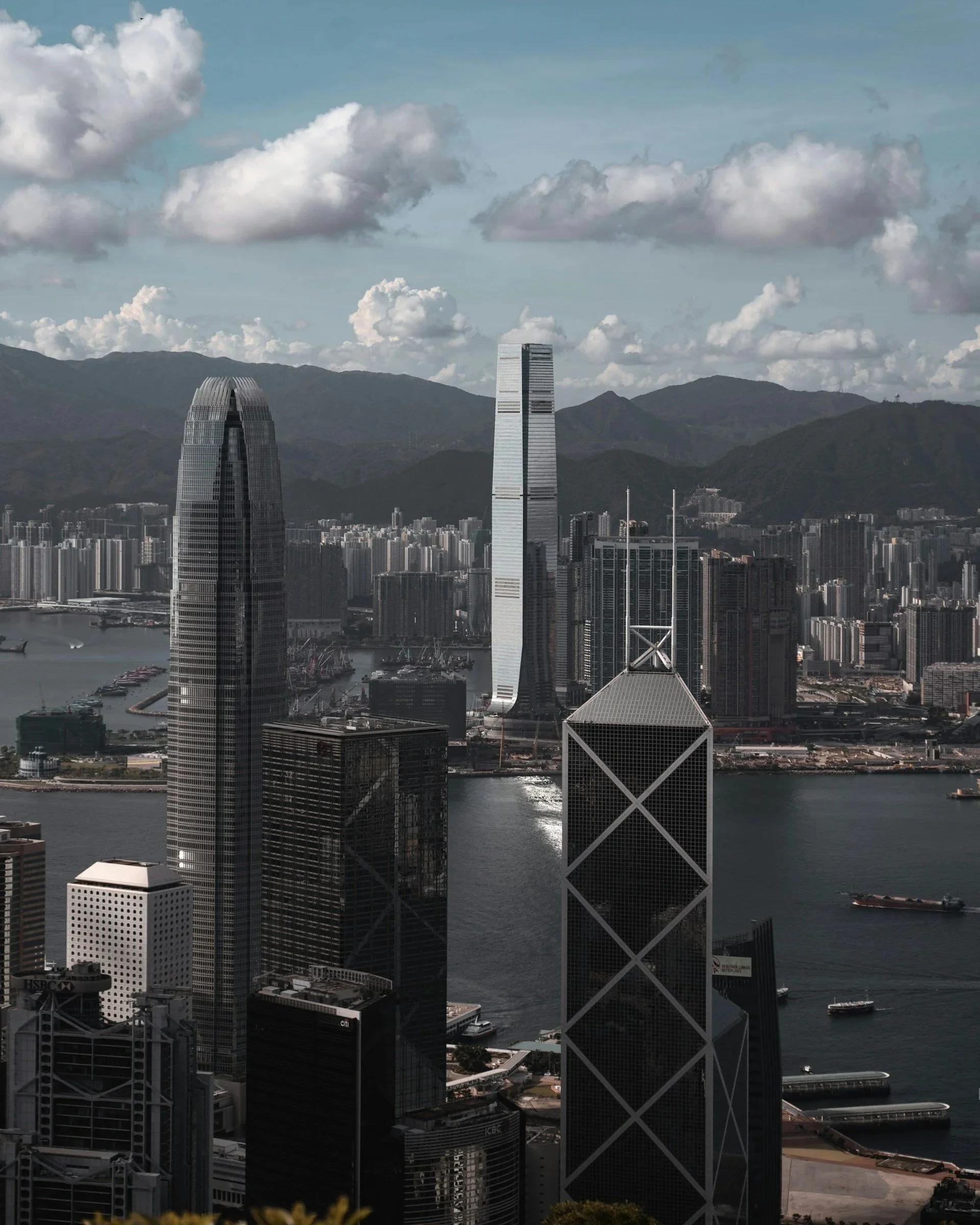 A city skyline with tall skyscrapers and mountains in the background, viewed over a body of water with boats. The tallest building is a modern high-rise with a distinctive white and black facade.