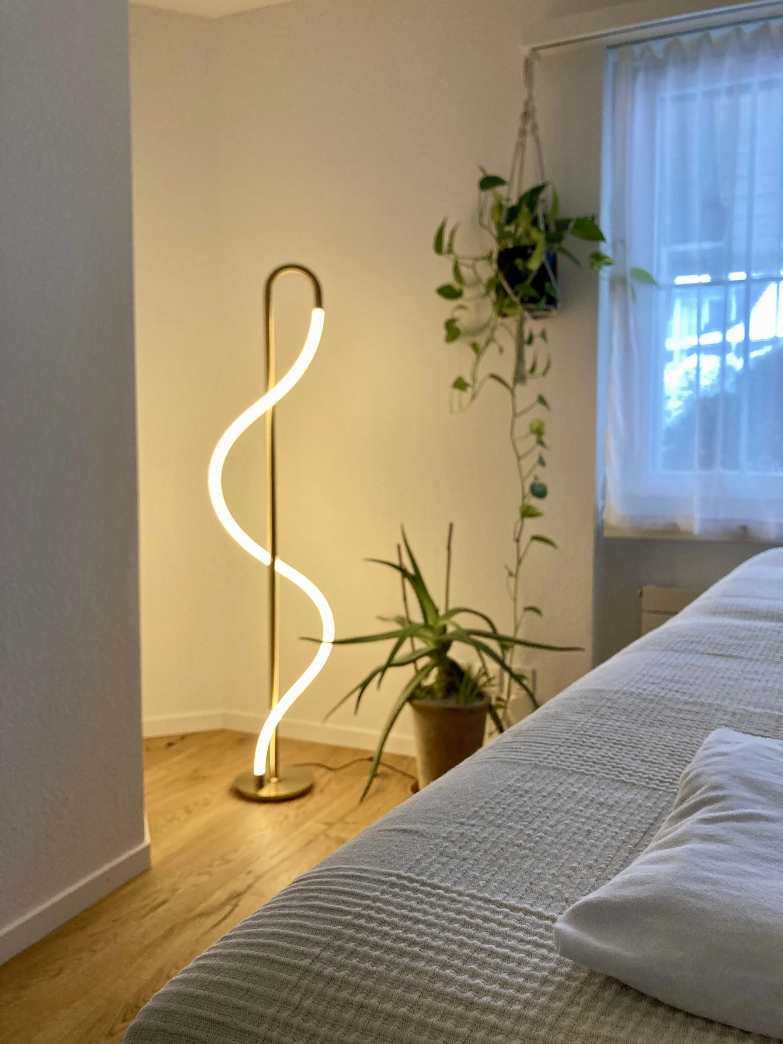 Ein modernes Stehlampe mit spiralförmiger Leuchte im Wohnzimmer neben Topfpflanzen und einem Bett, im Hintergrund ein Fenster mit weißen Vorhängen.