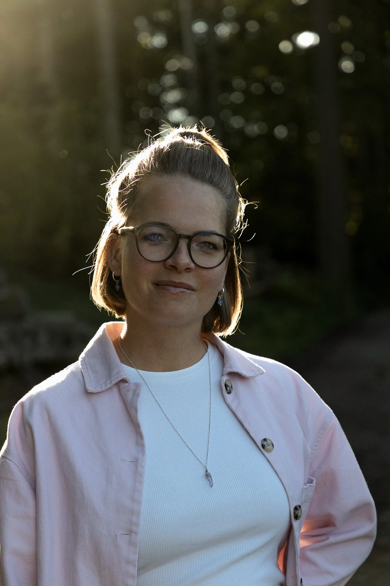 Junge Frau mit Brille und kurzem Haar, trägt ein weisses Oberteil und eine rosa Jacke, steht im Sonnenlicht im Freien, im Hintergrund sind Bäume und ein Pfad sichtbar.