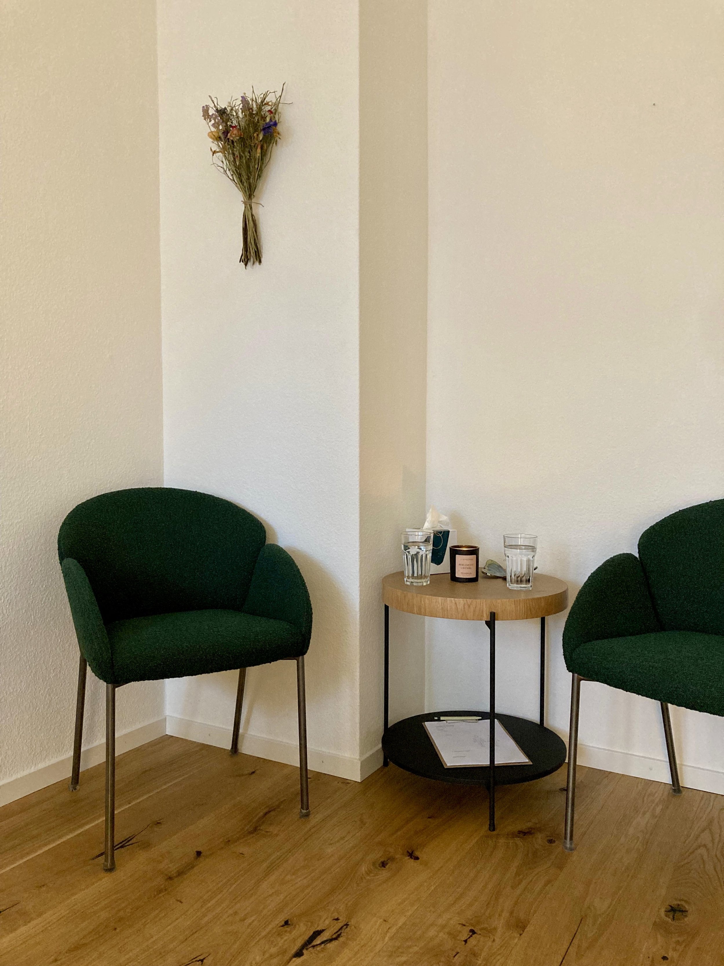 Zwei grüne Sessel, ein kleiner runder Tisch mit zwei Gläsern Wasser, einer Kerze und einem Paket Taschentücher, an einer weißen Wand mit einer Trockenblumen-Bündel an der Wand angebracht.