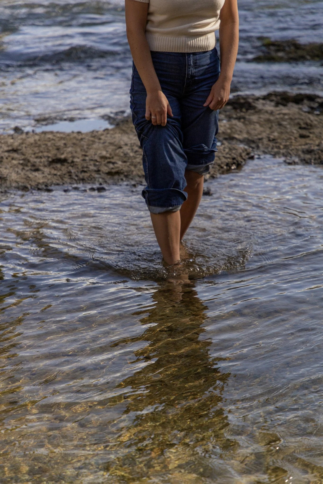 Person, die barfuß im Wasser an einem Strand läuft, mit Rollkragenpullover und Jeans, die die Hosenbeine hochgekrempelt hat.