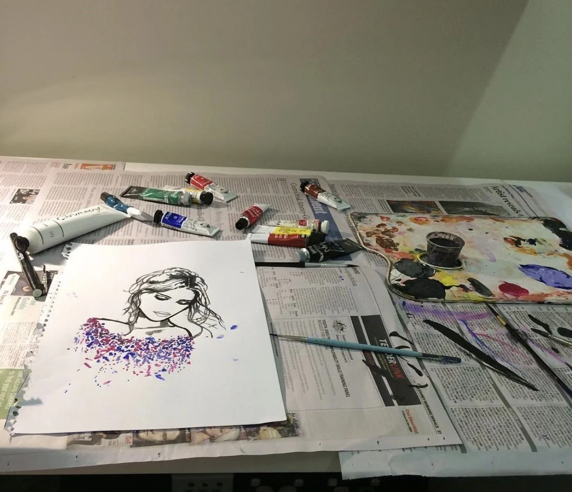 Art supplies including paint tubes, brushes, and a palette with mixed colors on a covered table, with a completed ink and watercolor portrait of a woman on a sheet of paper.
