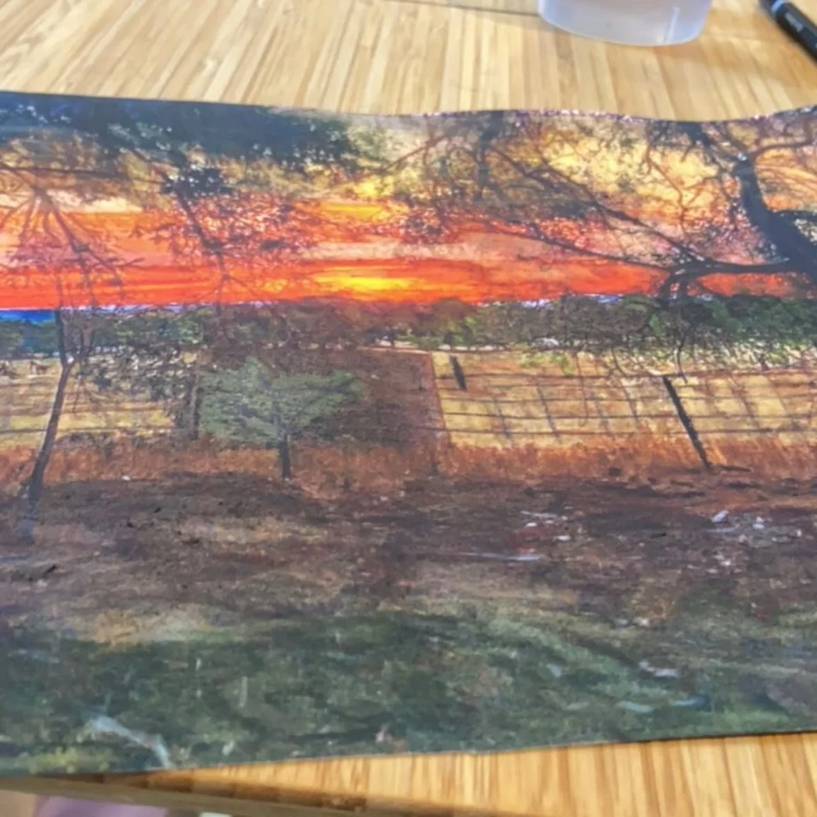 A landscape painting of a sunset over a farm with trees, fields, and a brick structure, on a wooden table.