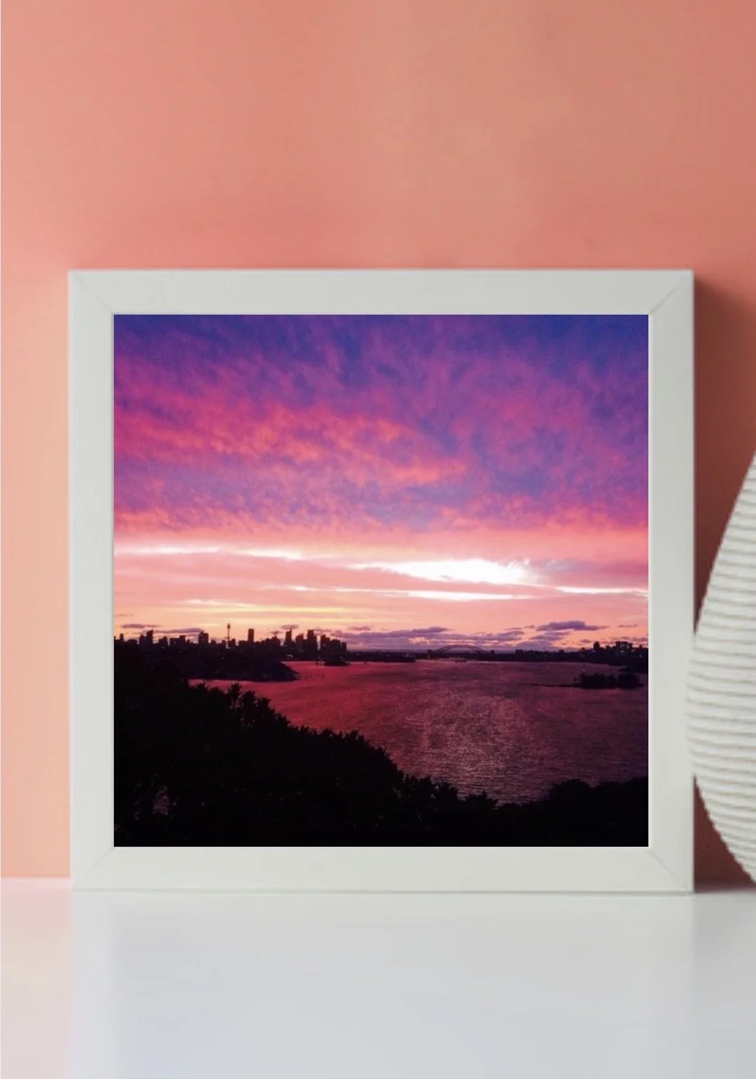 Framed photograph of a colorful sunset or sunrise with pink and purple clouds over a river and city skyline