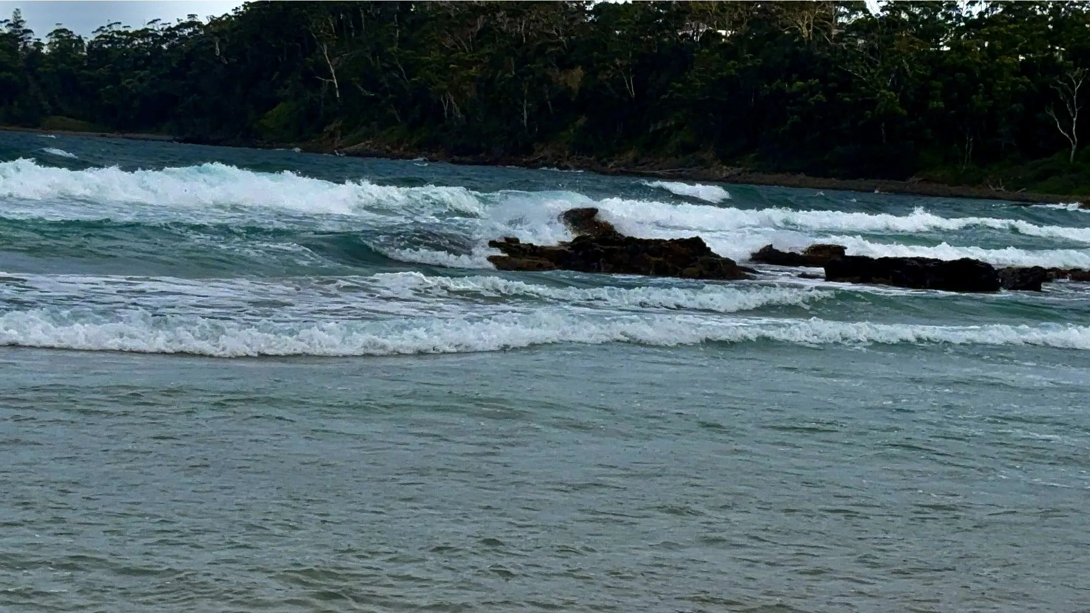 Ocean waves crashing onto a beach with rocks and a forested coastline in the background.
