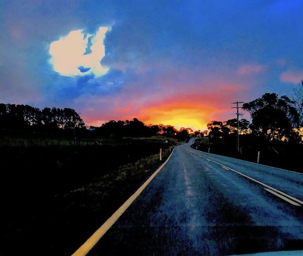 A country road at sunset with a colorful sky, clouds, and trees along the sides.