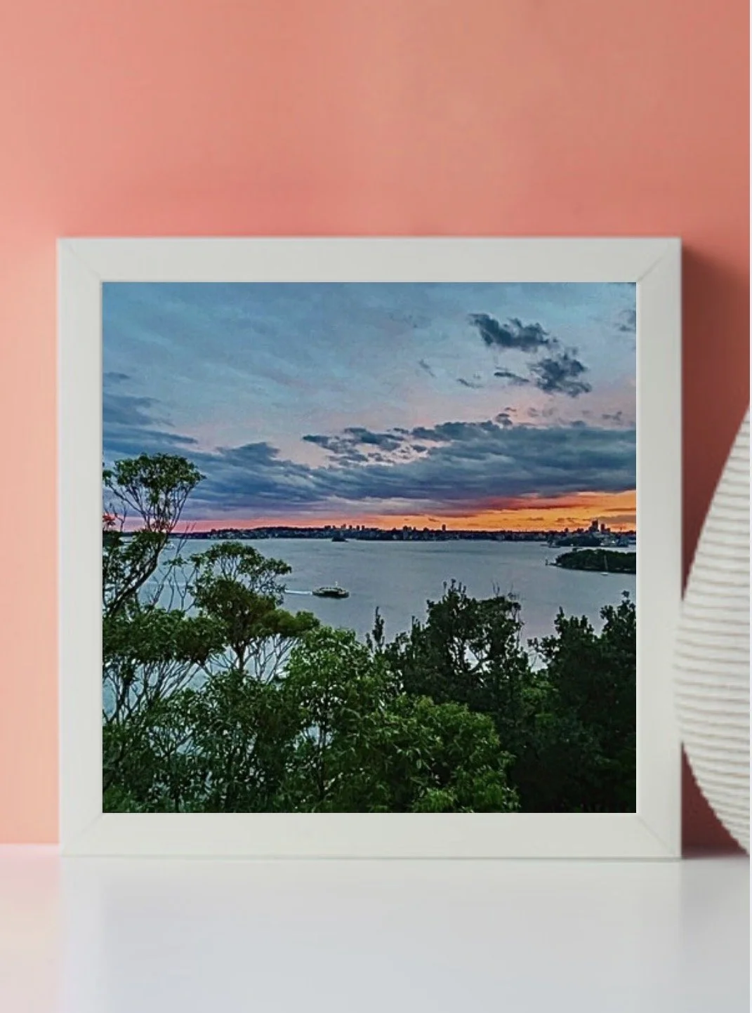 A framed landscape photograph of a sunset over a river, with trees in the foreground and a city skyline in the distance, set against a pink wall.