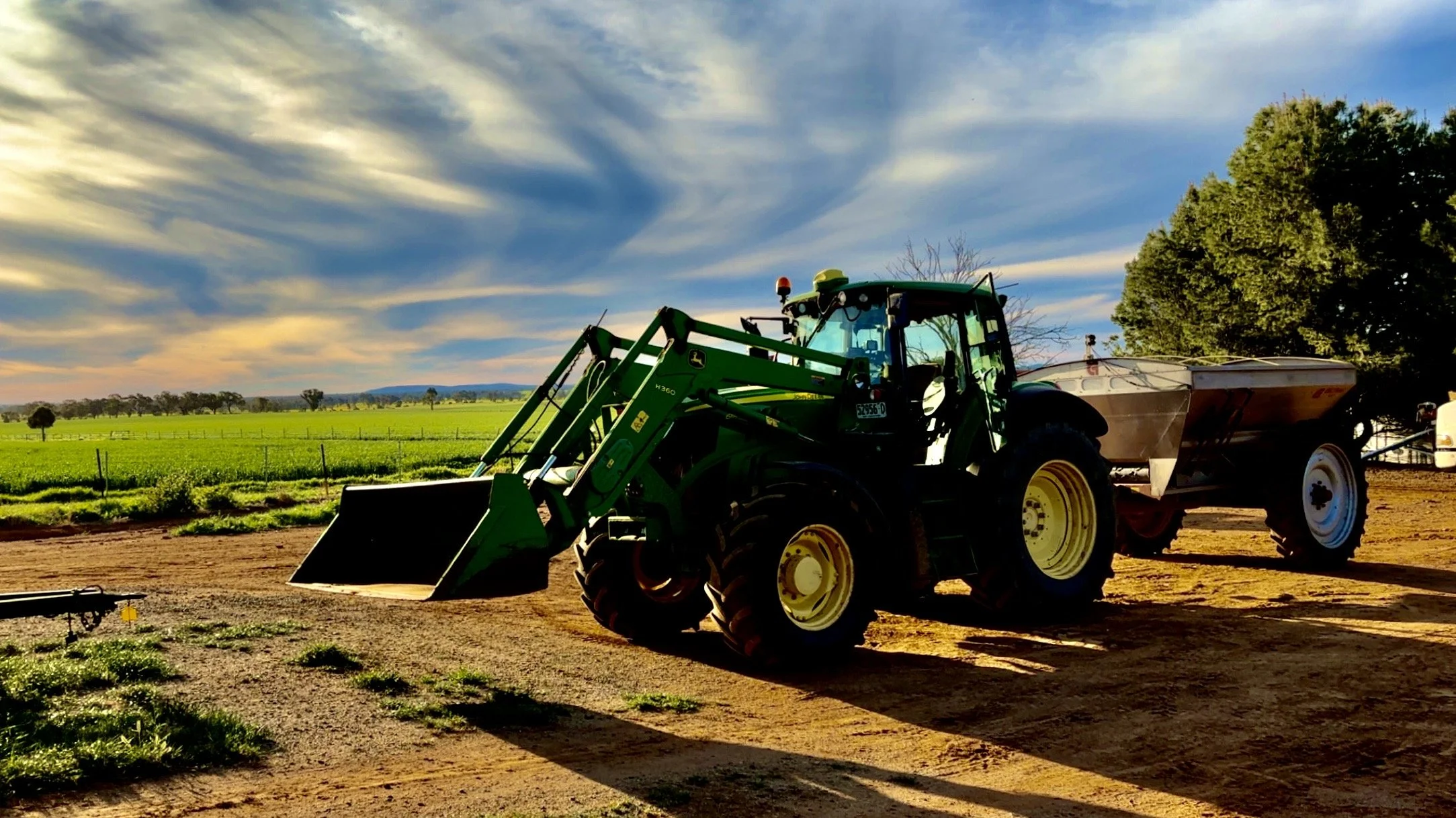 Green tractor with front loader and a trailer in a rural farmland area under a partly cloudy sky, with fields and trees in the background.