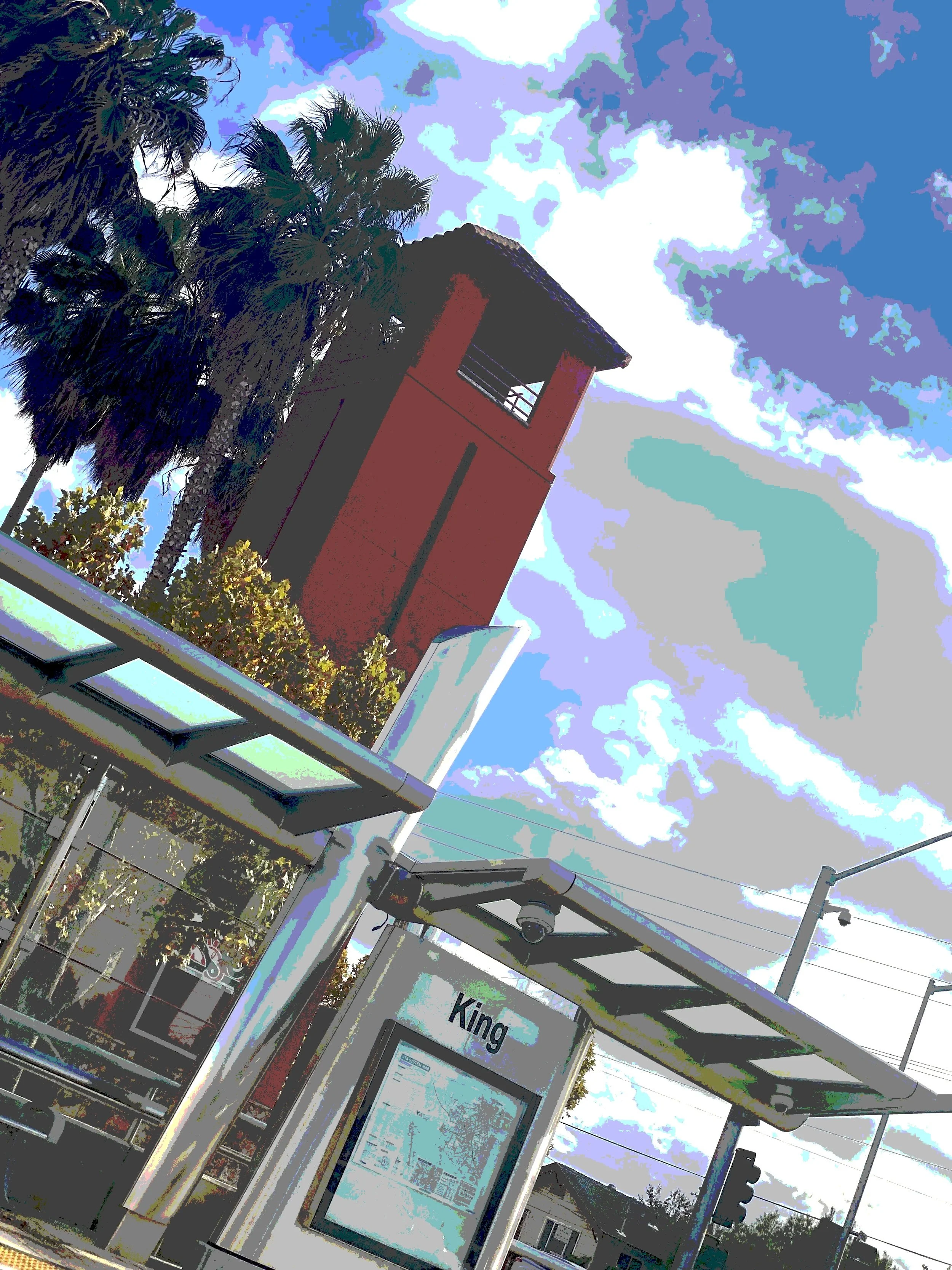 Street scene featuring a bus stop with an informational sign, a traffic light, and a tall red clock tower with palm trees and a partly cloudy sky in the background.