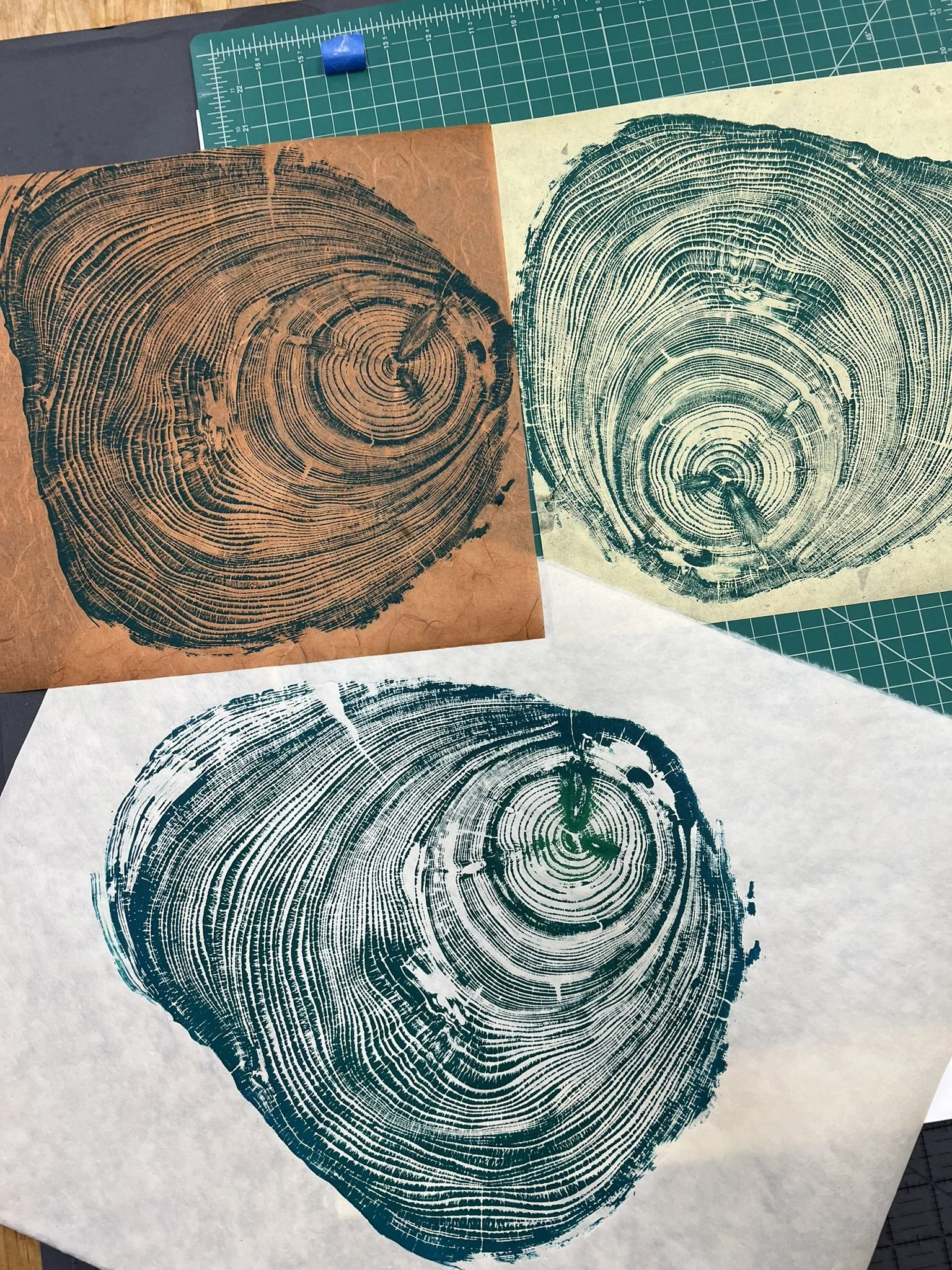 Three prints of tree trunk cross-sections, showing growth rings, on different colored papers, with a green cutting mat and a recycling ruler in the background.