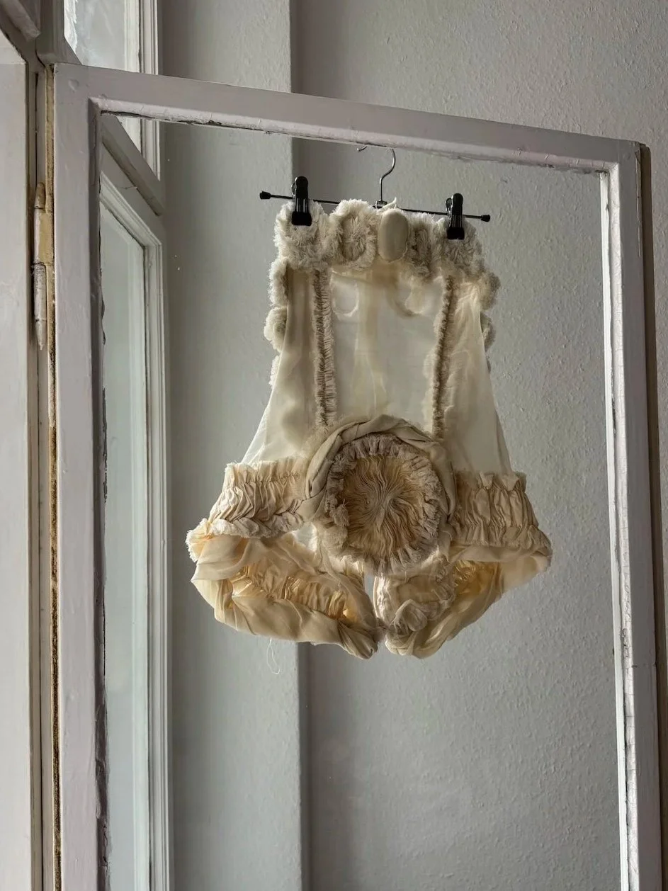 Erinnerungsstück-Janiejean-Couture-Ein weißer Baby-Overall mit Fellbesatz hängt an einem schwarzen Kleiderbügel vor einem Fenster