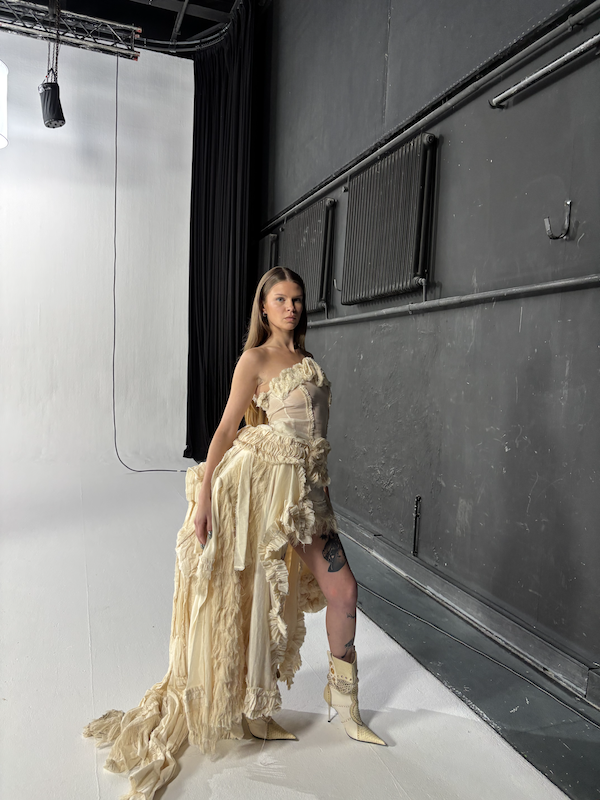 Frau posiert vor dunkler Wand in einem Studio, trägt ein extravagant gelbes Kleid mit Rüschen und hohen Schuhen.