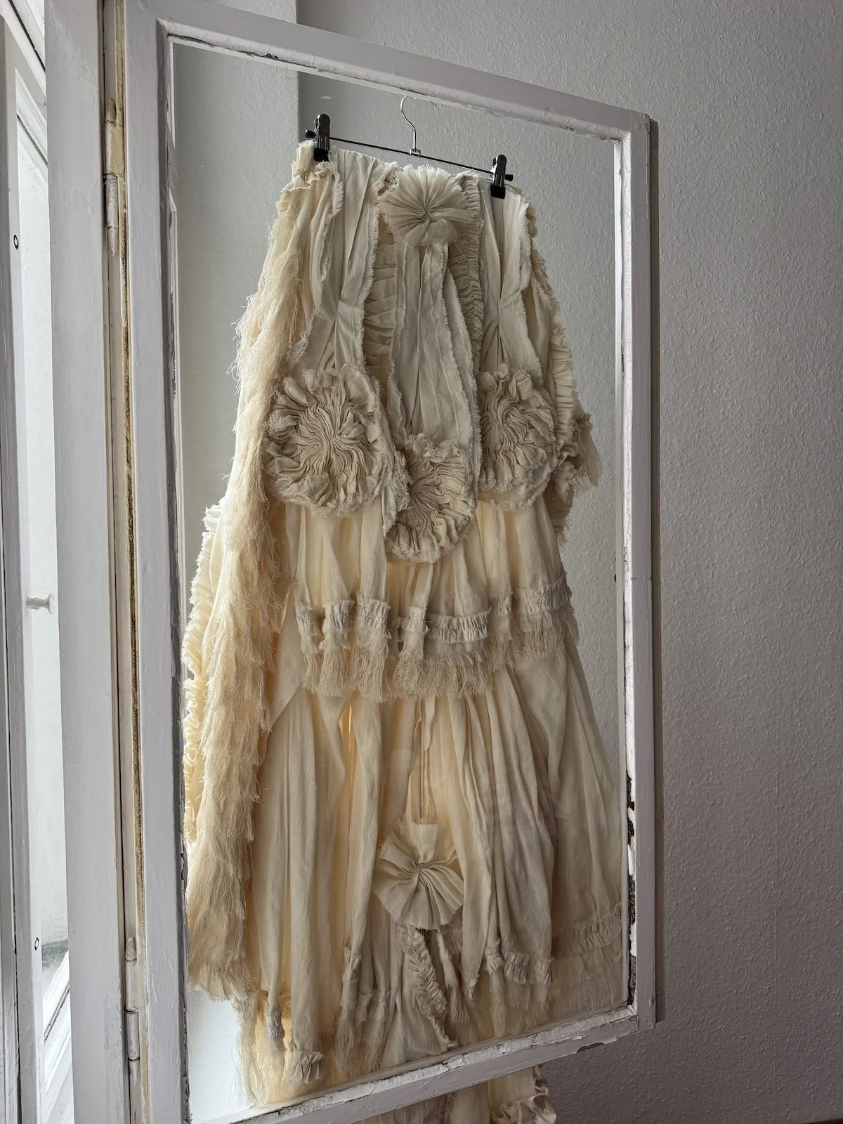 Erinnerungsstück-Janiejean-Couture-Ein cremefarbener, zerfetzter und geraffter Auftritt eines Kleidungsstücks, das im Spiegel hängt.