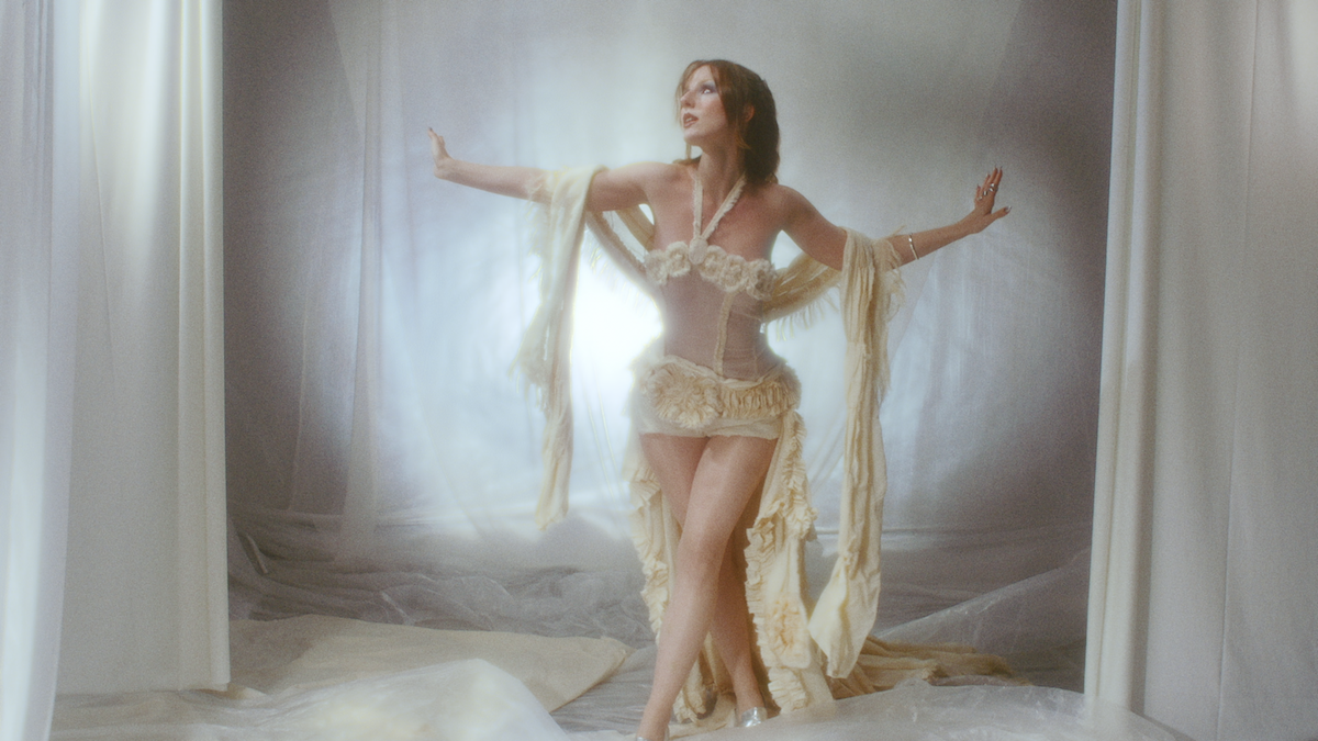 Eine Frau in einem auffälligen, cremefarbenen Kostüm posiert vor einem Hintergrund mit weißen Vorhängen, mit einem dramatischen Lichteffekt hinter ihr.