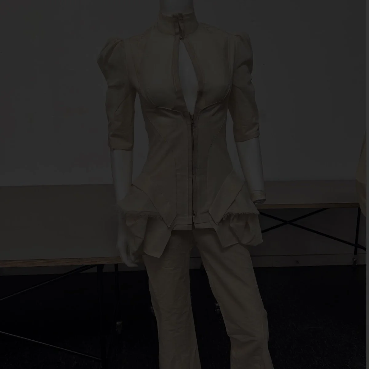 Identity-Janiejean Ein modisches, beige Outfit auf einem Mannequin, bestehend aus einer langen Bluse mit hohen Schultern und einer weißen Hose.