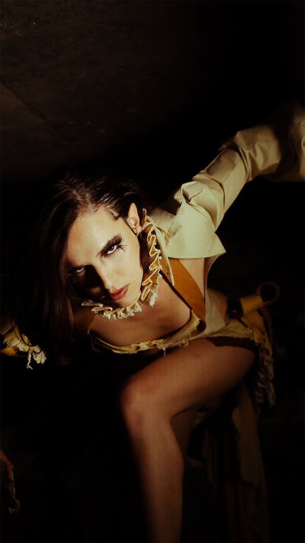 Eine junge Frau mit dunklen Haaren, im Lichtlicht betrachtet, in einer Khakifarbigen Jacke und einer cremefarbenen Bluse, in einer dunklen Umgebung fotografiert.