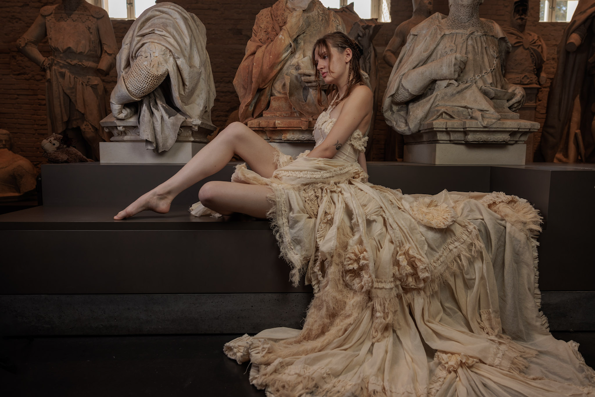 Erinnerungsstück-Janiejean-Couture-Junge Frau in einem aufwändigen, cremefarbenen Vintage-Kleid sitzt auf einer schwarzen Plattform in einem Raum mit Statuen und Skulpturen aus Marmor.