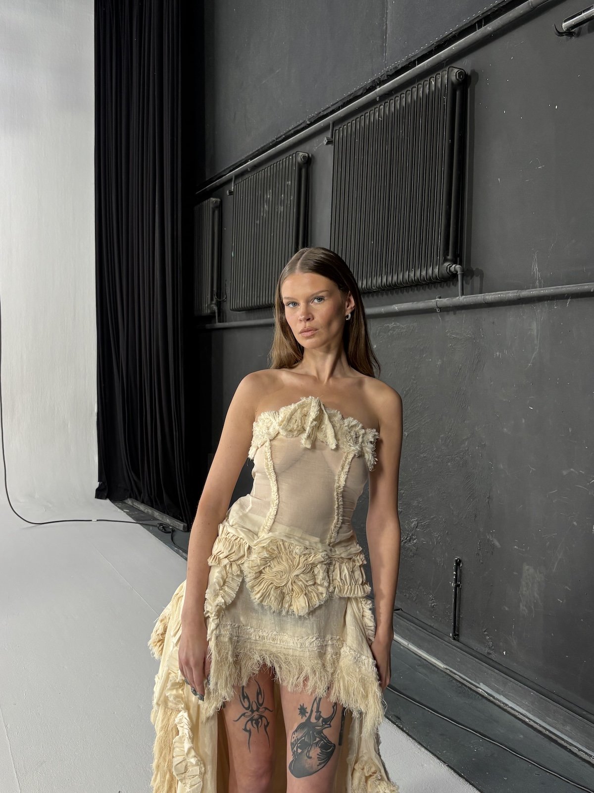 Eine junge Frau trägt ein ausgefallenes, cremefarbenes Kleid mit Rüschen und Textur im Studio mit dunklen Wänden und schwarzen Heizkörpern.