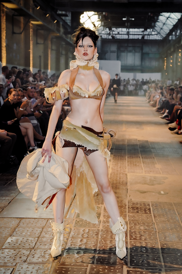 Model läuft auf Laufsteg während einer Modenschau, trägt ausgefallene, künstlerische Kleidung und Schuhe, im Hintergrund sind Zuschauer und eine industrienähnliche Kulisse zu sehen.