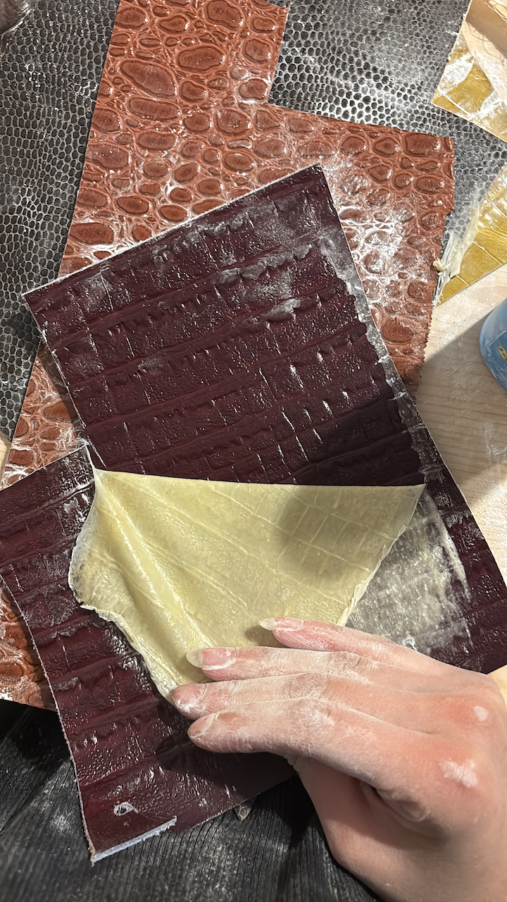 Janiejean-Latex-Textile-Surface, Hand, die dünnen, vierschichtigen Blätter aus Pastaproduktion auf dem Tisch rollt, mit verschiedenen Farbmustern in Schwarz, Braun, und Cremetönen.