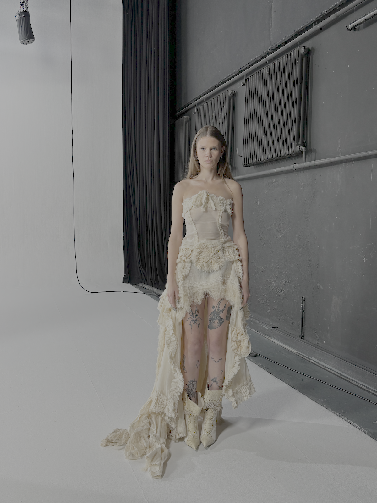 Eine junge Frau in einem außergewöhnlichen, zerzausten, cremefarbenen Kleid mit Tattoos an den Beinen steht auf einem Fotoset mit dunklem Hintergrund und schwarzen Vorhängen.