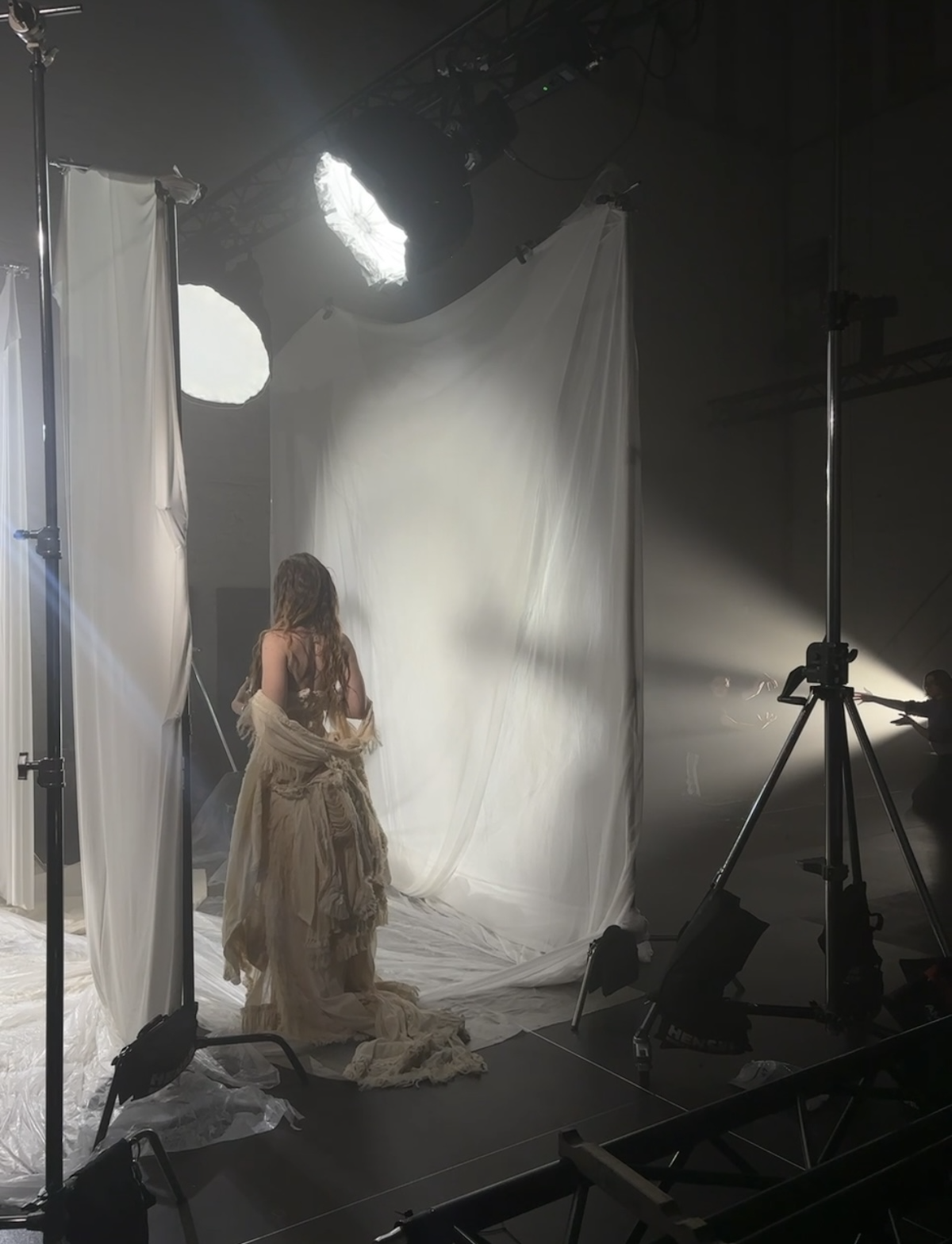 Eine Frau in einem langen, zerlumpten Kleid steht vor einem weißen Stoffhintergrund in einem Fotostudio, umgeben von Studiobeleuchtung und Kamerastativen.