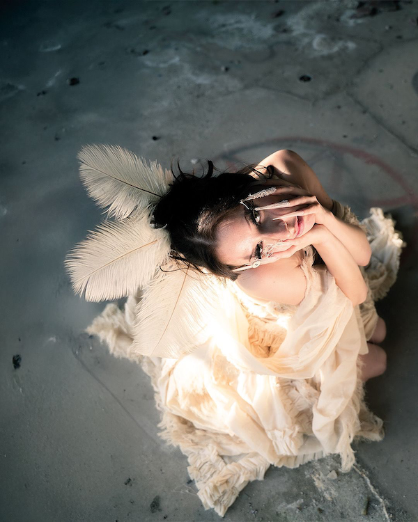 Erinnerungsstück-Janiejean-Couture-Eine Frau mit dunklem Haar in einem weißen Kleid und großen Federn im Haar sitzt auf einem Boden, schaut nach oben und berührt ihr Gesicht.