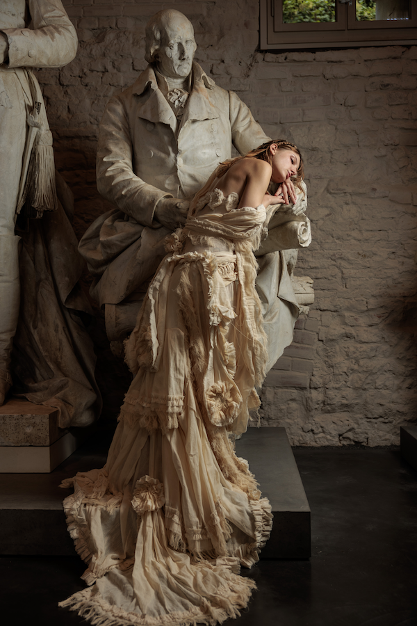 Erinnerungsstück-Janiejean-Couture-Eine junge Frau in einem alten, zerfledderten Kleid liegt auf einem Bronzebrunnen, umgeben von antiken Skulpturen in einem Museum.